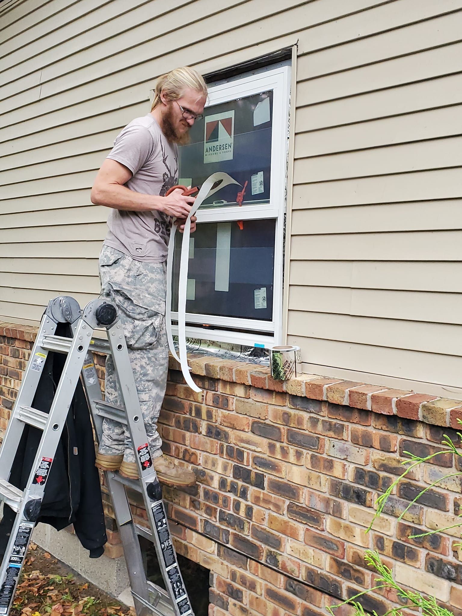 Person on a ladder painting a window frame on a brick-and-siding house exterior.