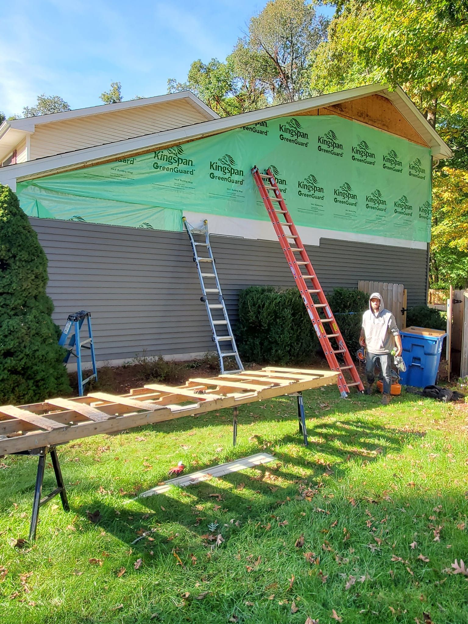 House exterior under repair with green tarp, two ladders, and workers in a yard