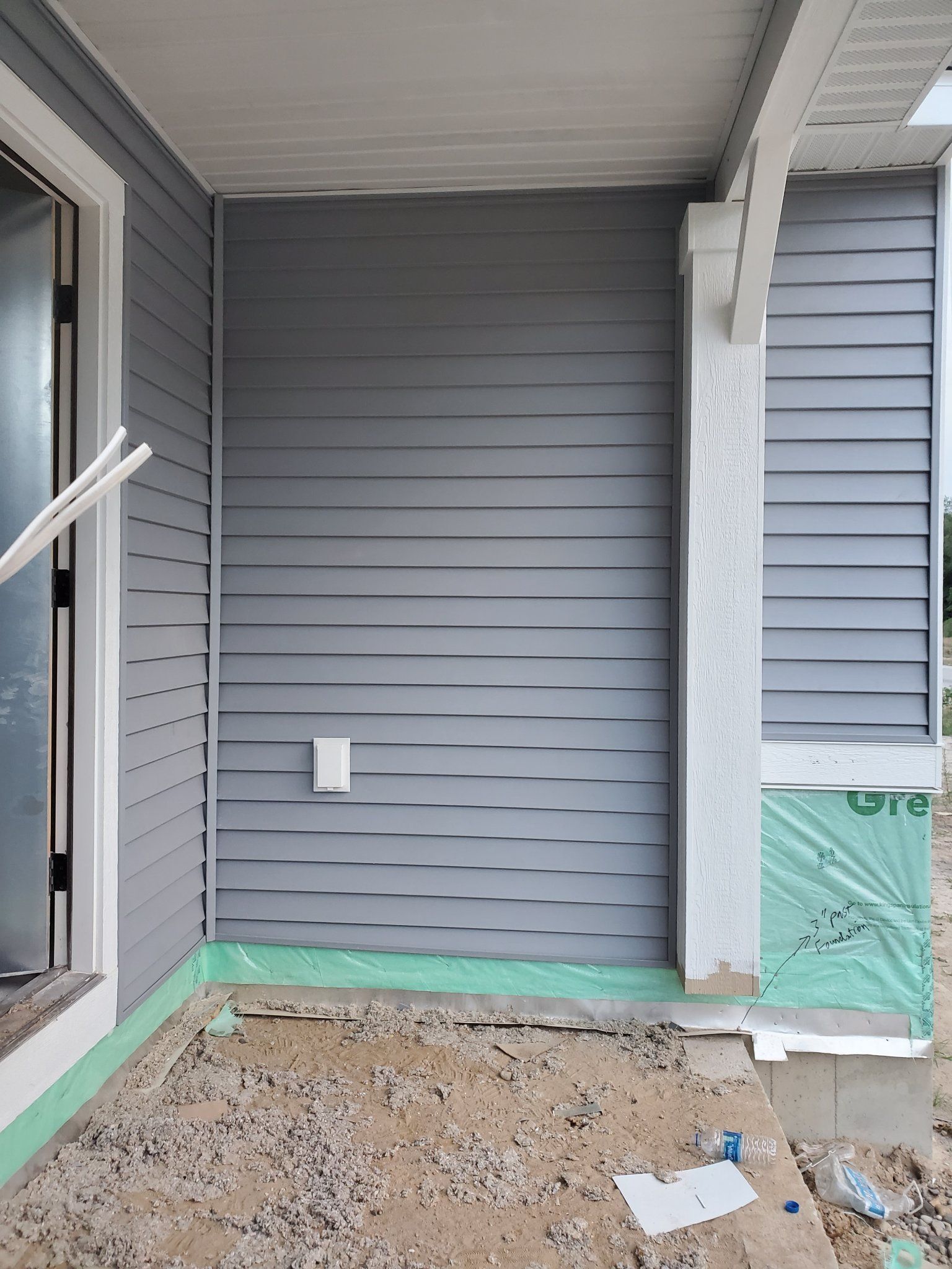 Front porch under construction with gray siding, concrete floor, and green protective wrap on the right side.
