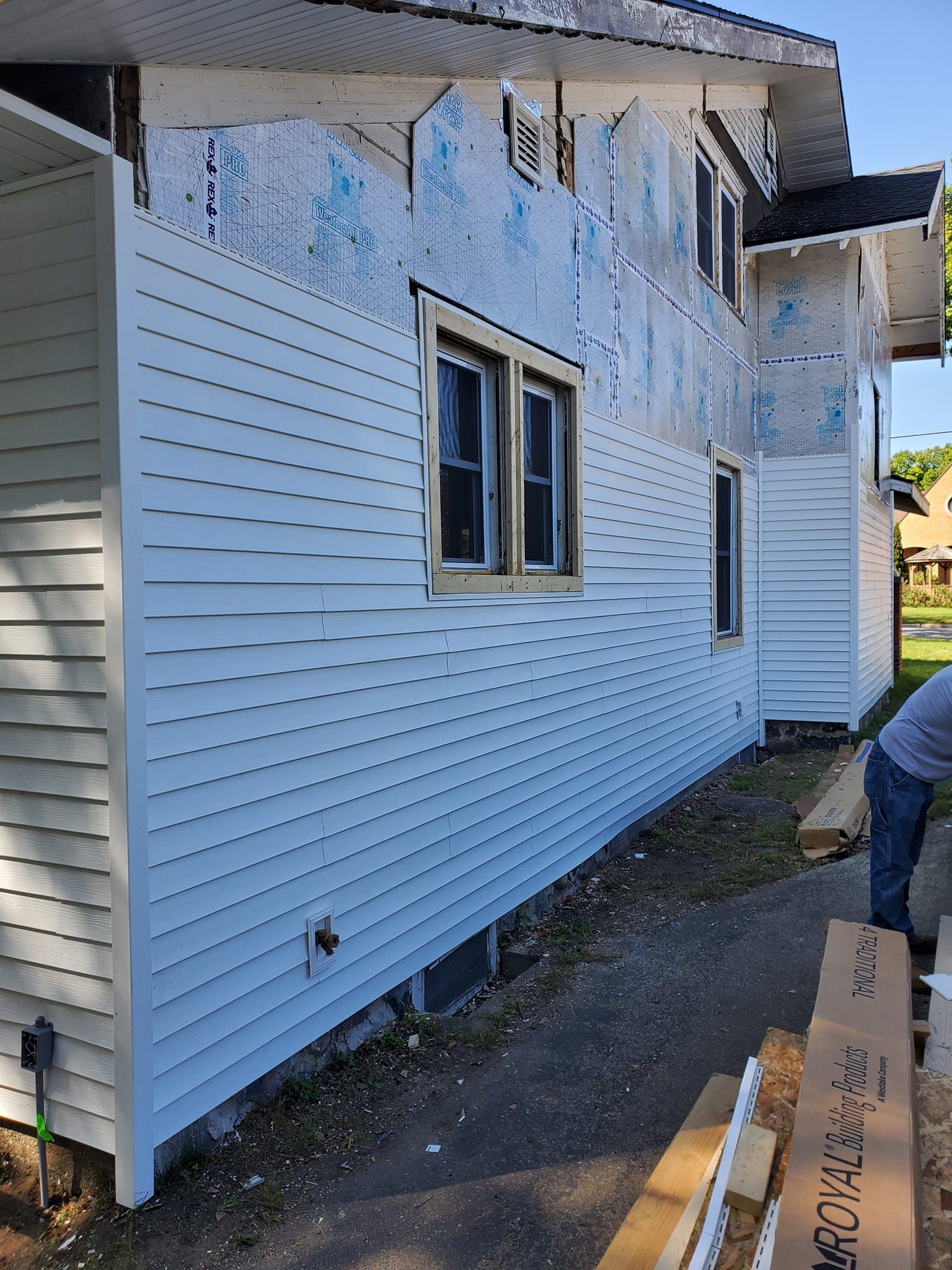 Two-story house with white siding under repair, peeling blue paint, and construction materials beside the wall.