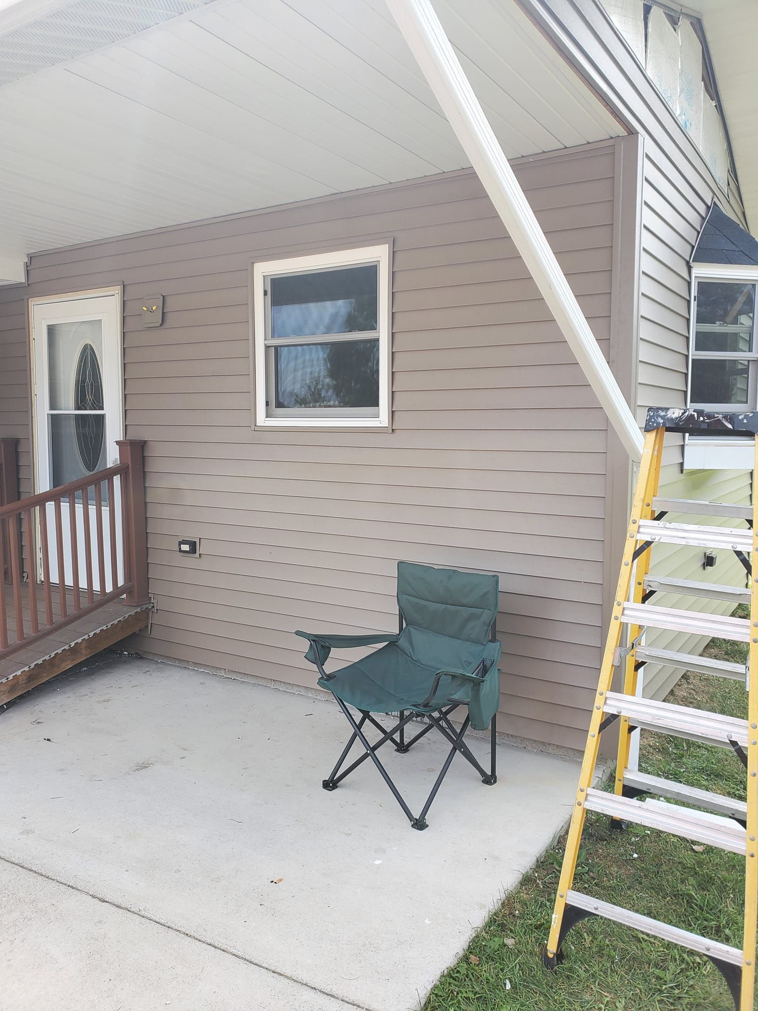 Covered porch with folding chair, window, and ladder leaning against the siding.