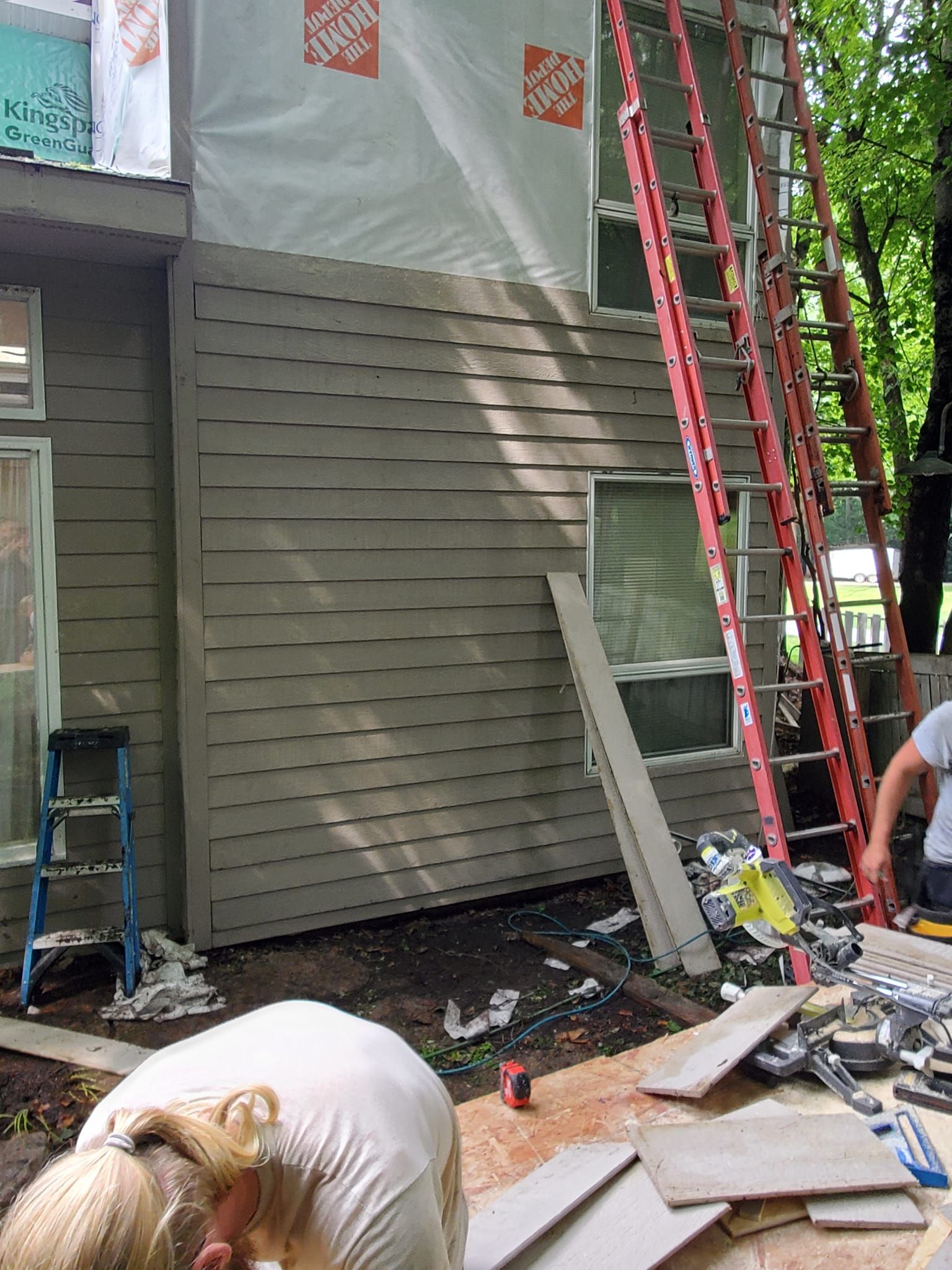 Workers and ladders outside a house under repair, with tools and debris on the ground
