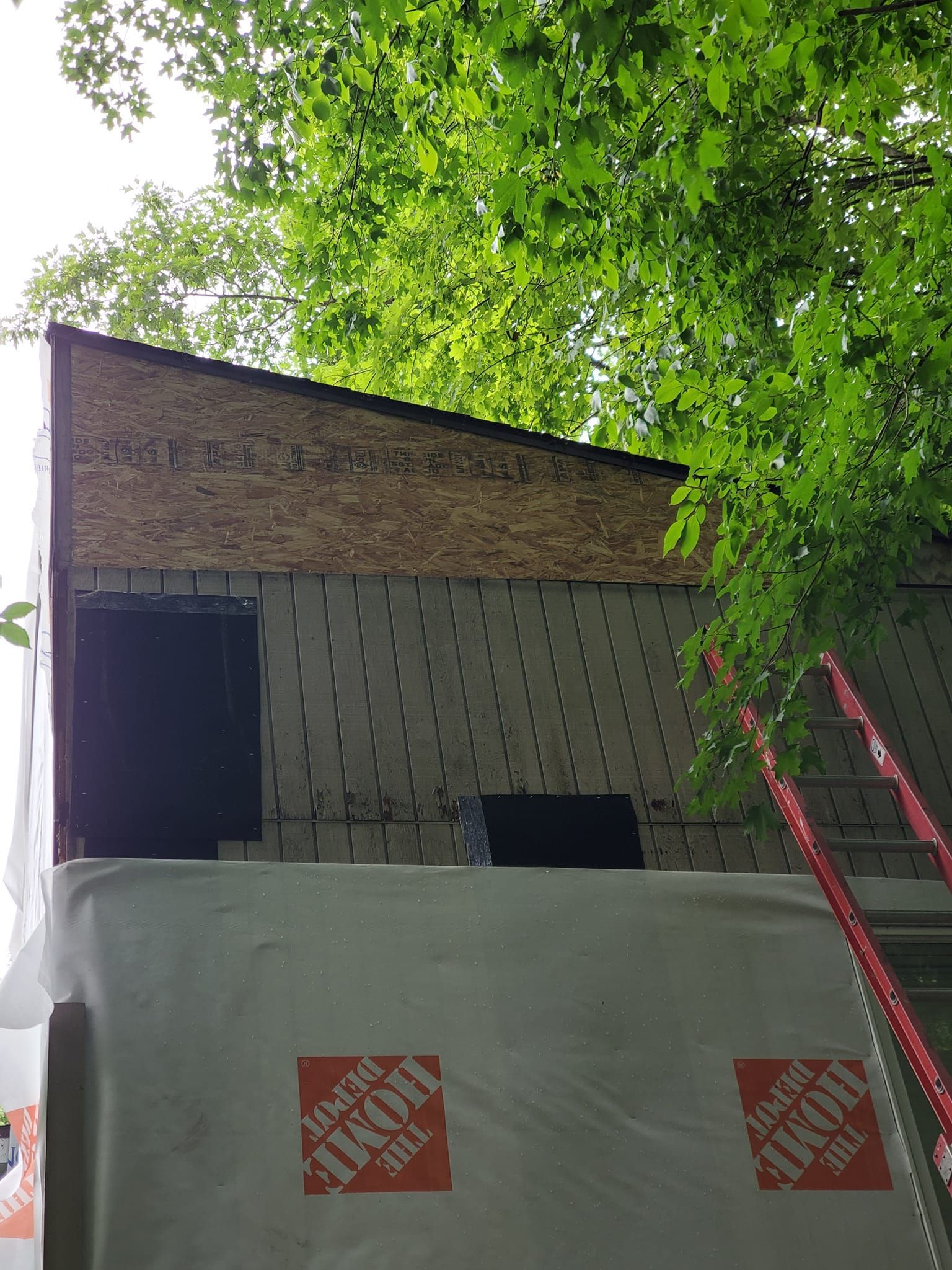 Partially built house with brick and siding, wrapped in white Tyvek, with red ladder and leafy trees overhead