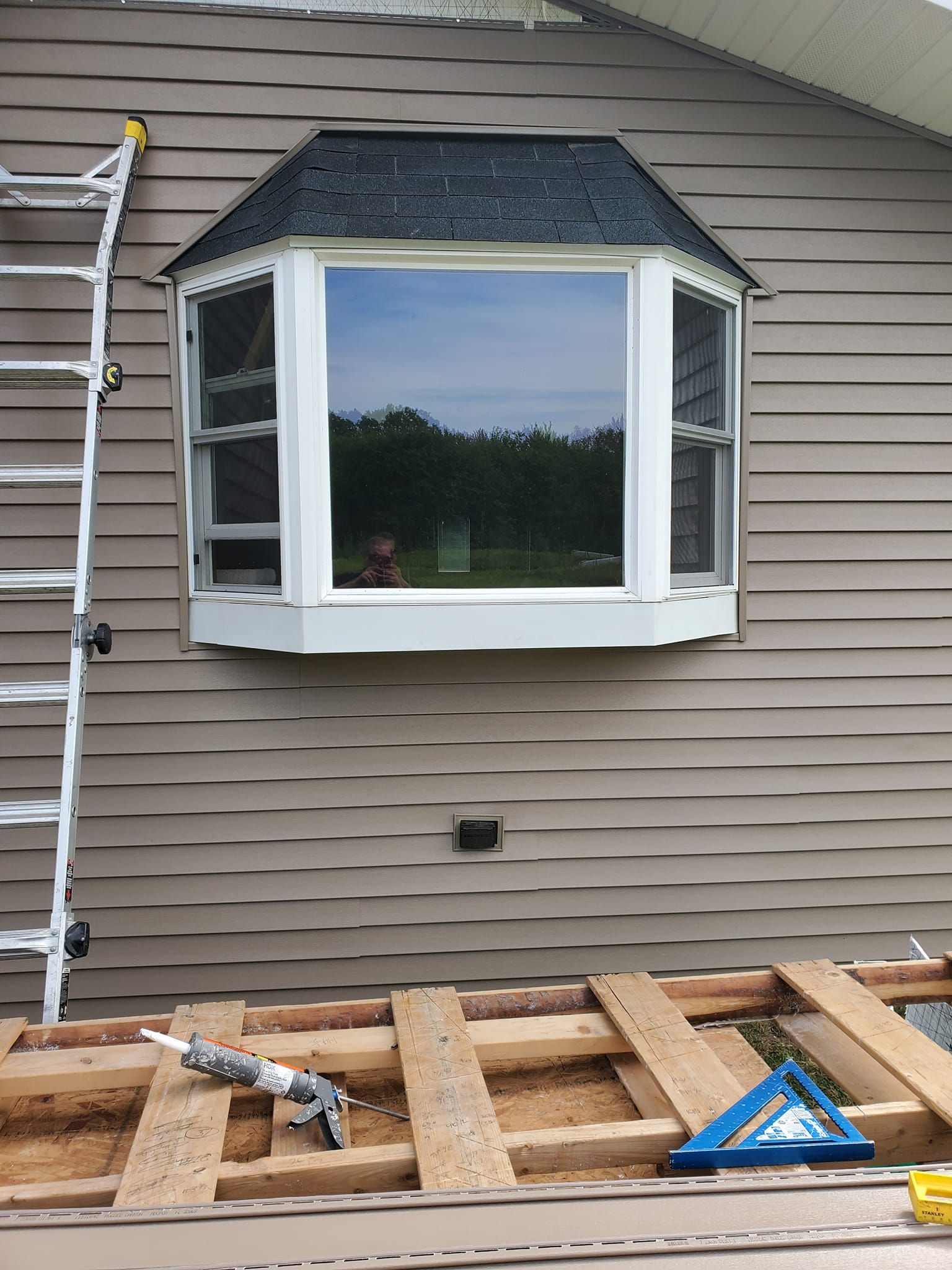 Bay window on tan siding, with roof shingles and construction materials on a wooden deck below.
