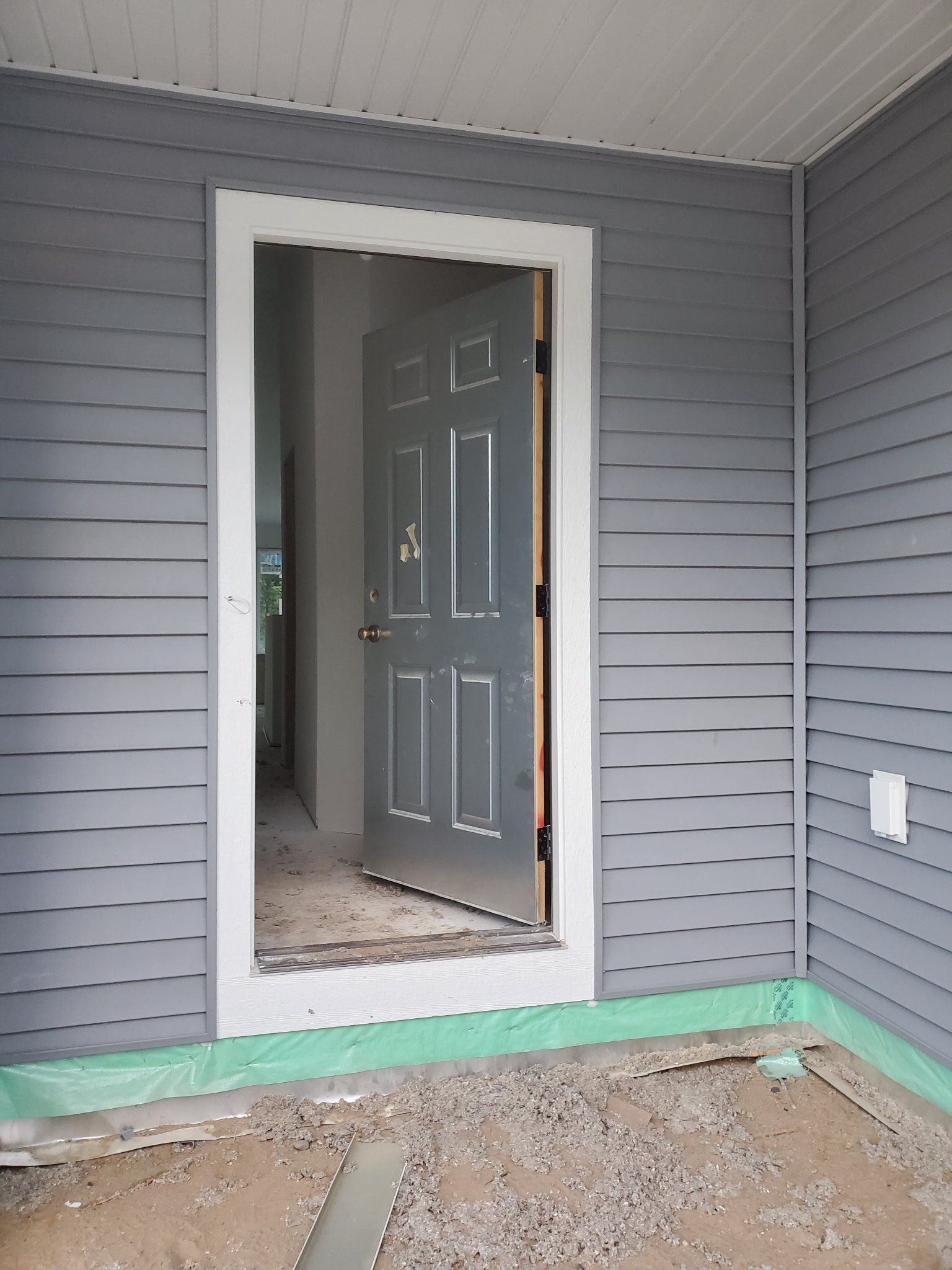 Front porch with gray siding, open doorway, and unfinished dirt ground at the entrance