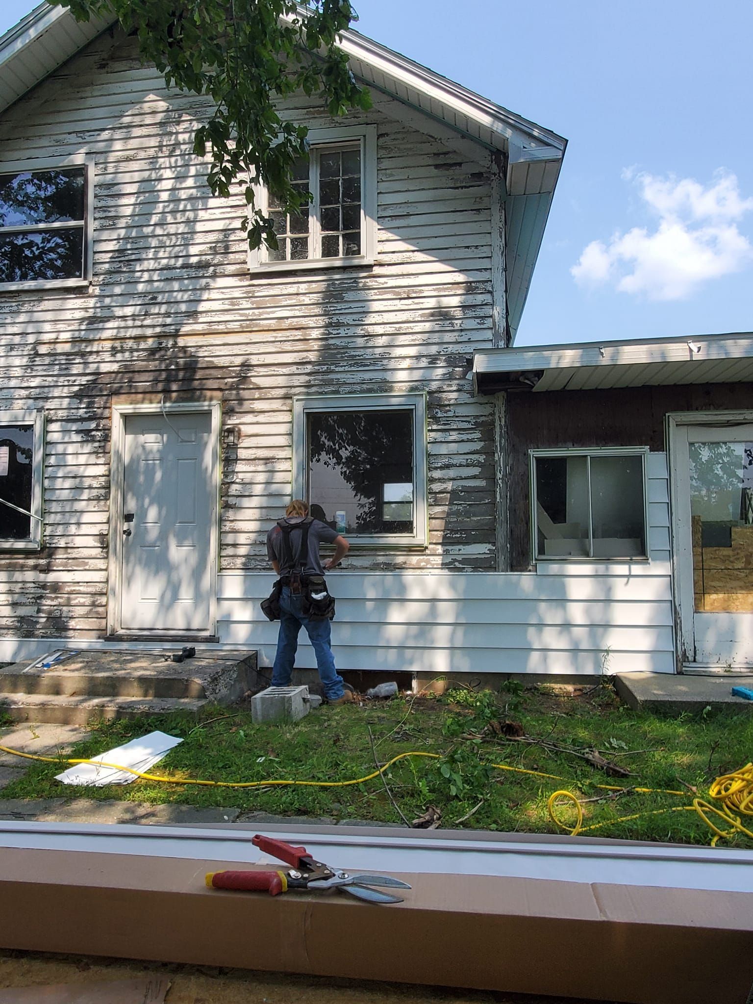 Person standing beside a weathered white house with boarded windows and a porch in daylight.