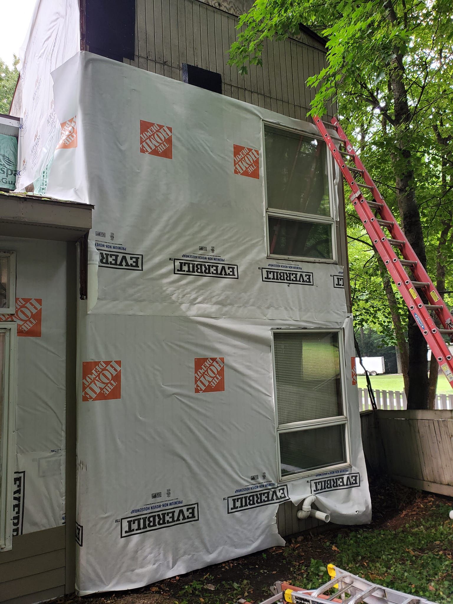Wrapped outdoor unit beside a building, with a red ladder and green tree overhead.