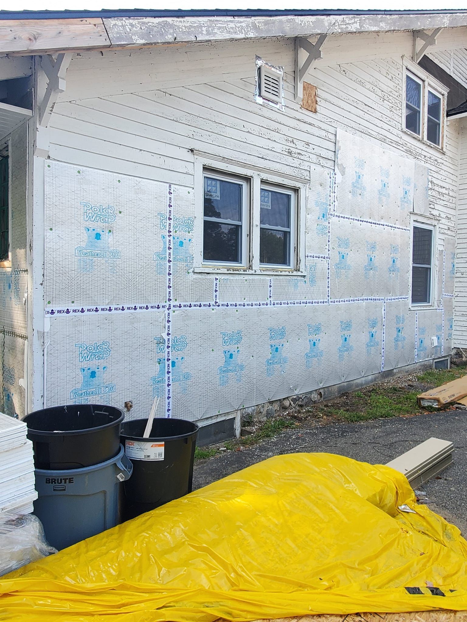House exterior under construction with yellow tarp, trash bins, and exposed insulation panels