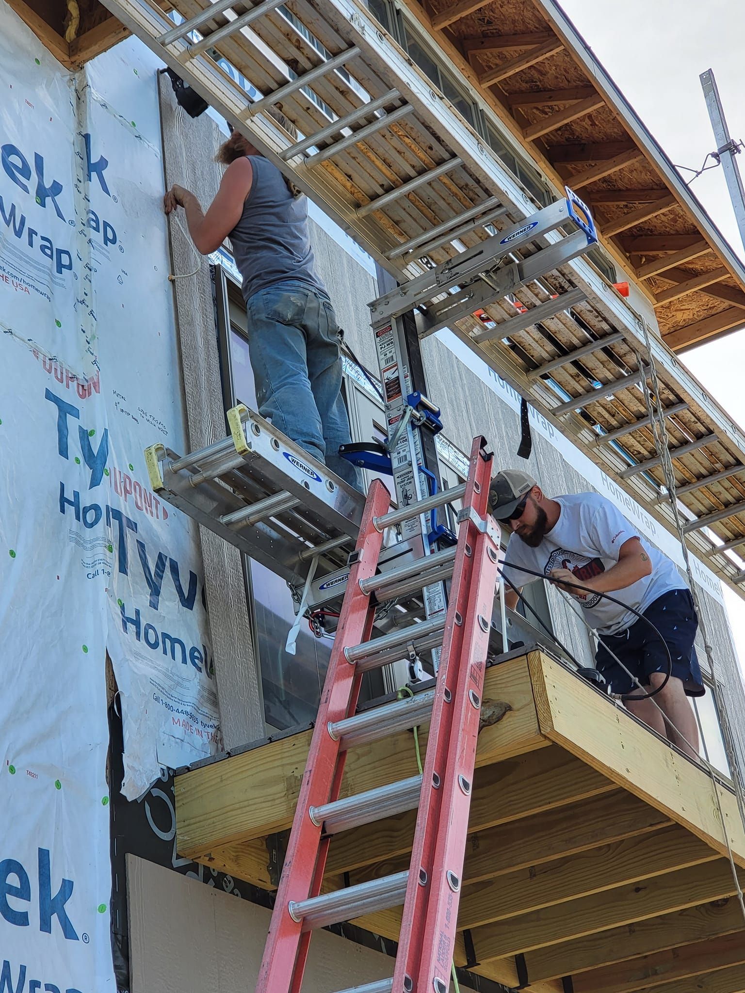 Two workers on ladders installing siding on a house exterior under construction