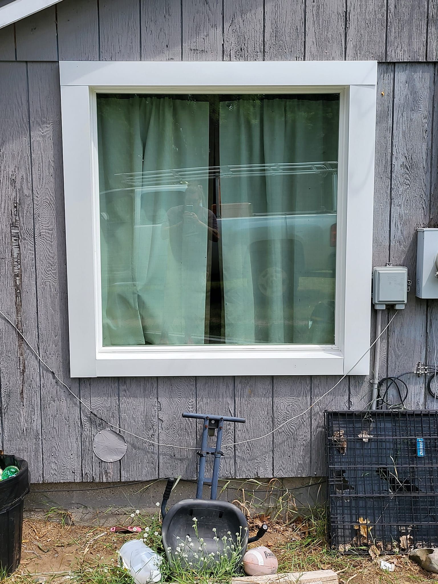 Window on weathered gray wall with white trim, pale green curtain, and yard tools and bins below