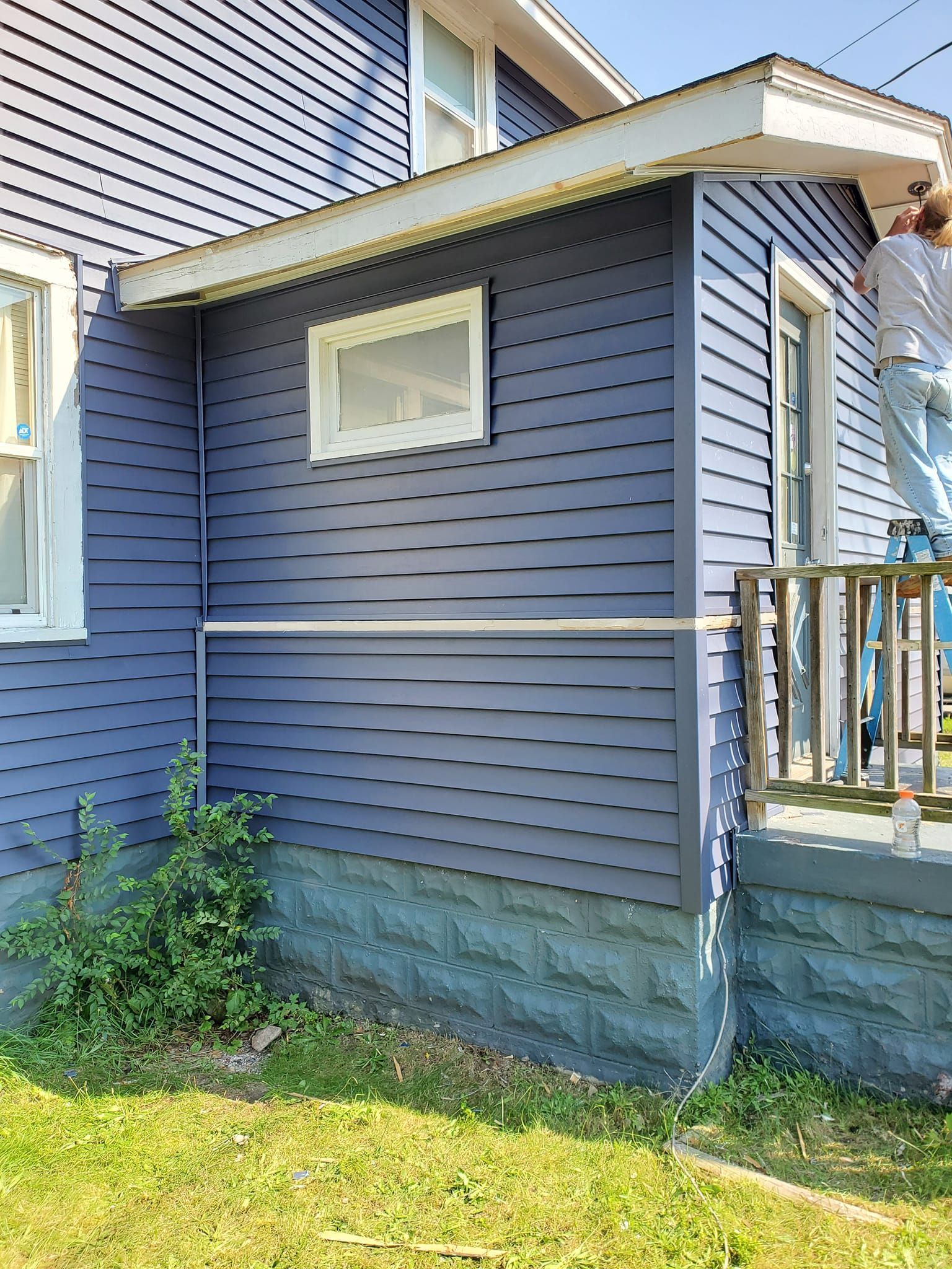 Blue-sided house exterior with a small window, porch railing, and green lawn in front