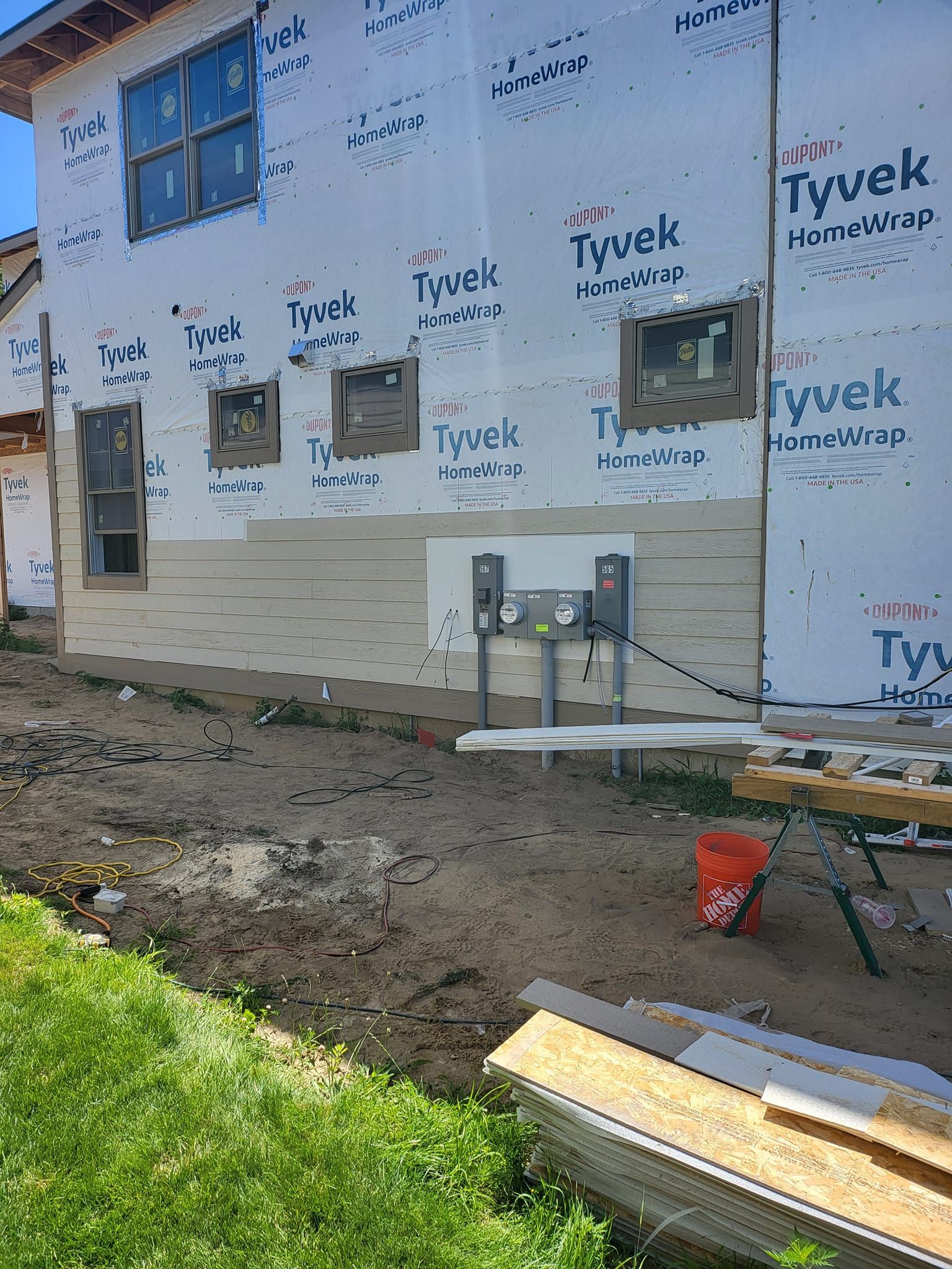 Exterior of a house under construction with Tyvek wrap, windows, dirt ground, and stacked lumber.