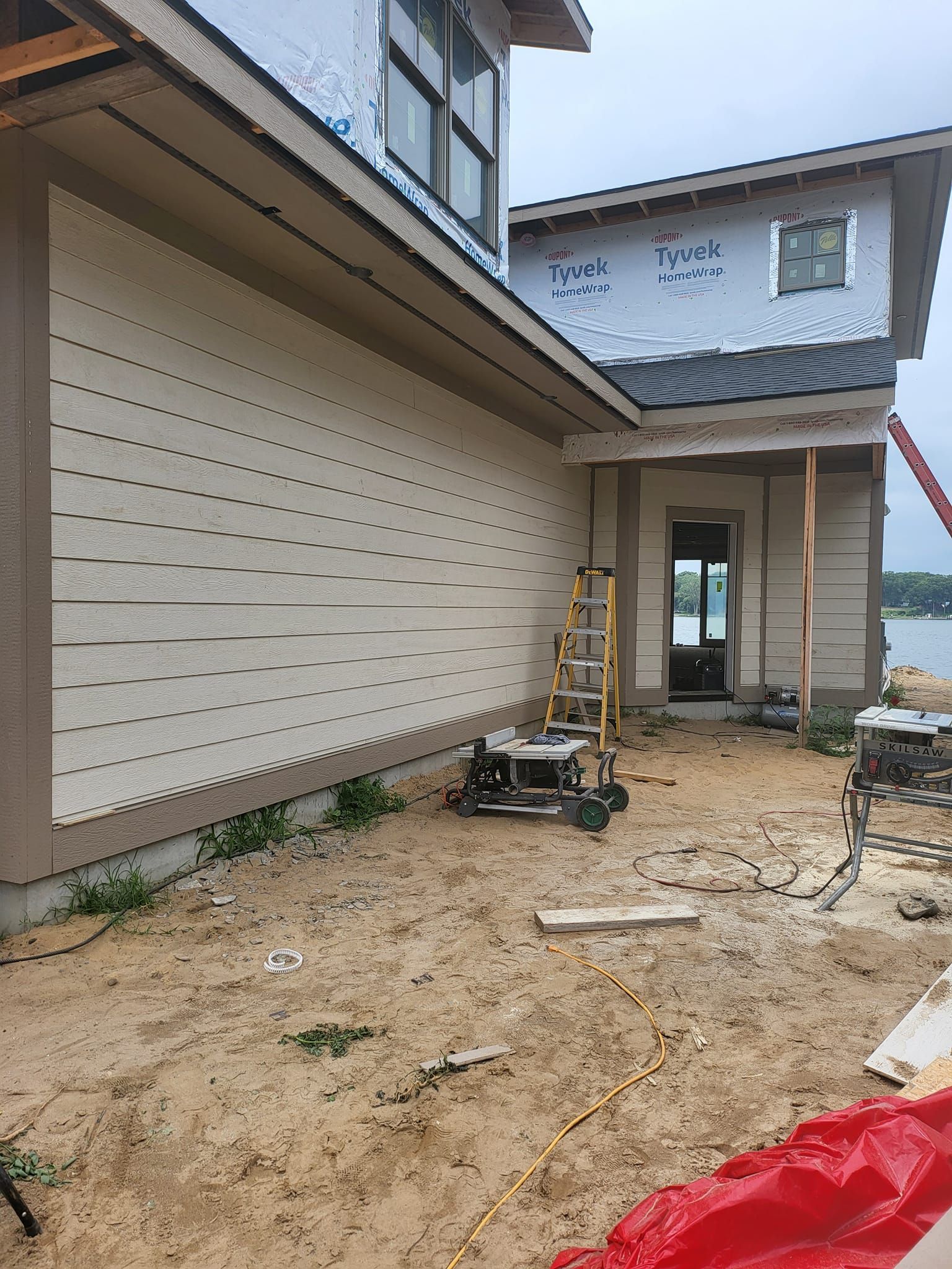 Exterior of a house under construction, with unfinished siding, dirt yard, and scattered tools.