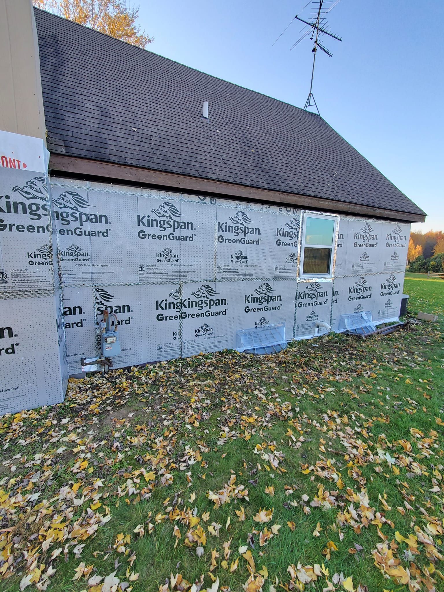 House under construction wrapped in Tyvek, with dark roof shingles and a small window, surrounded by fallen leaves