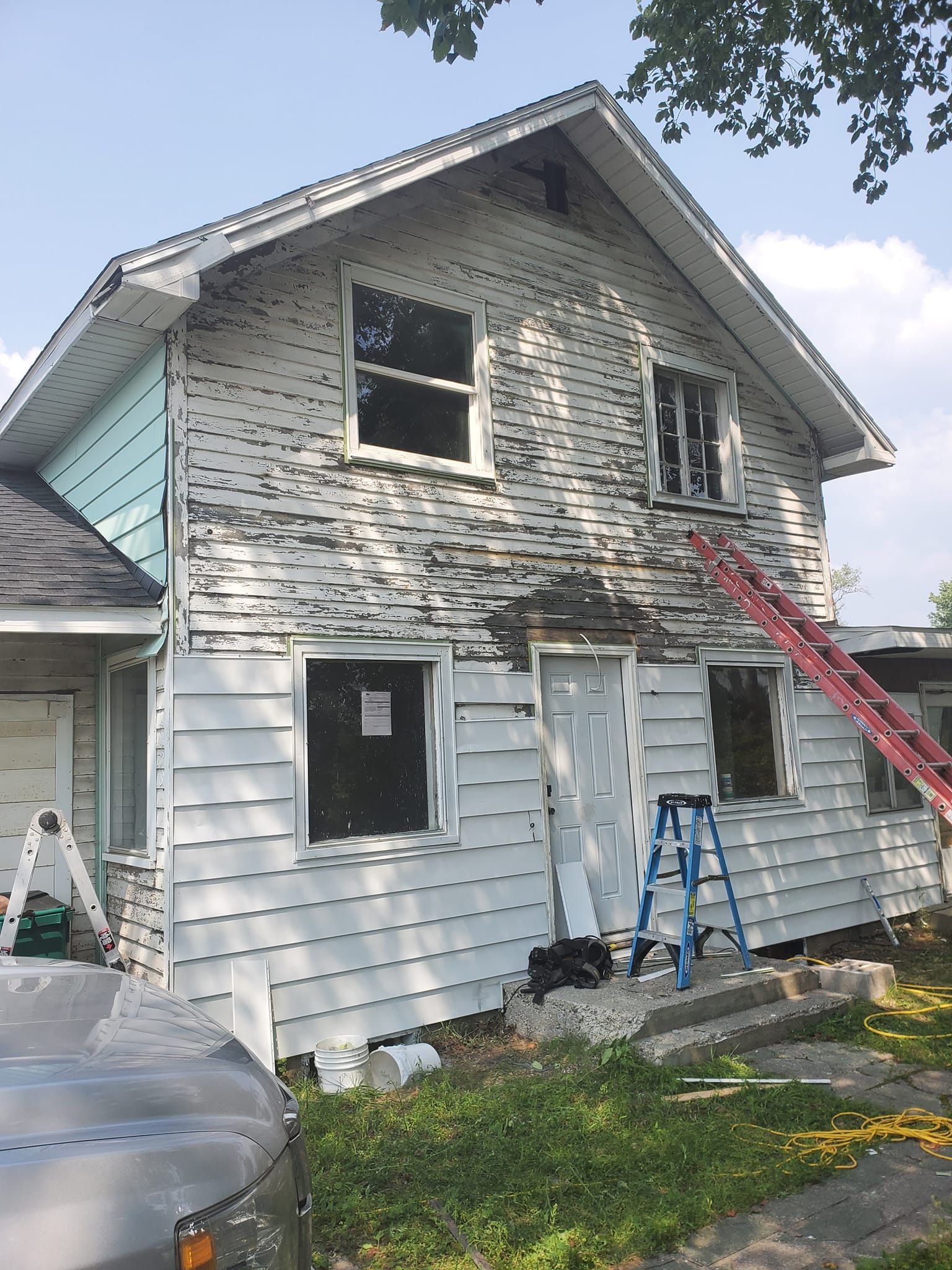 Two-story house with white siding and exposed upper wall, red ladder leaning against it, and tools below.