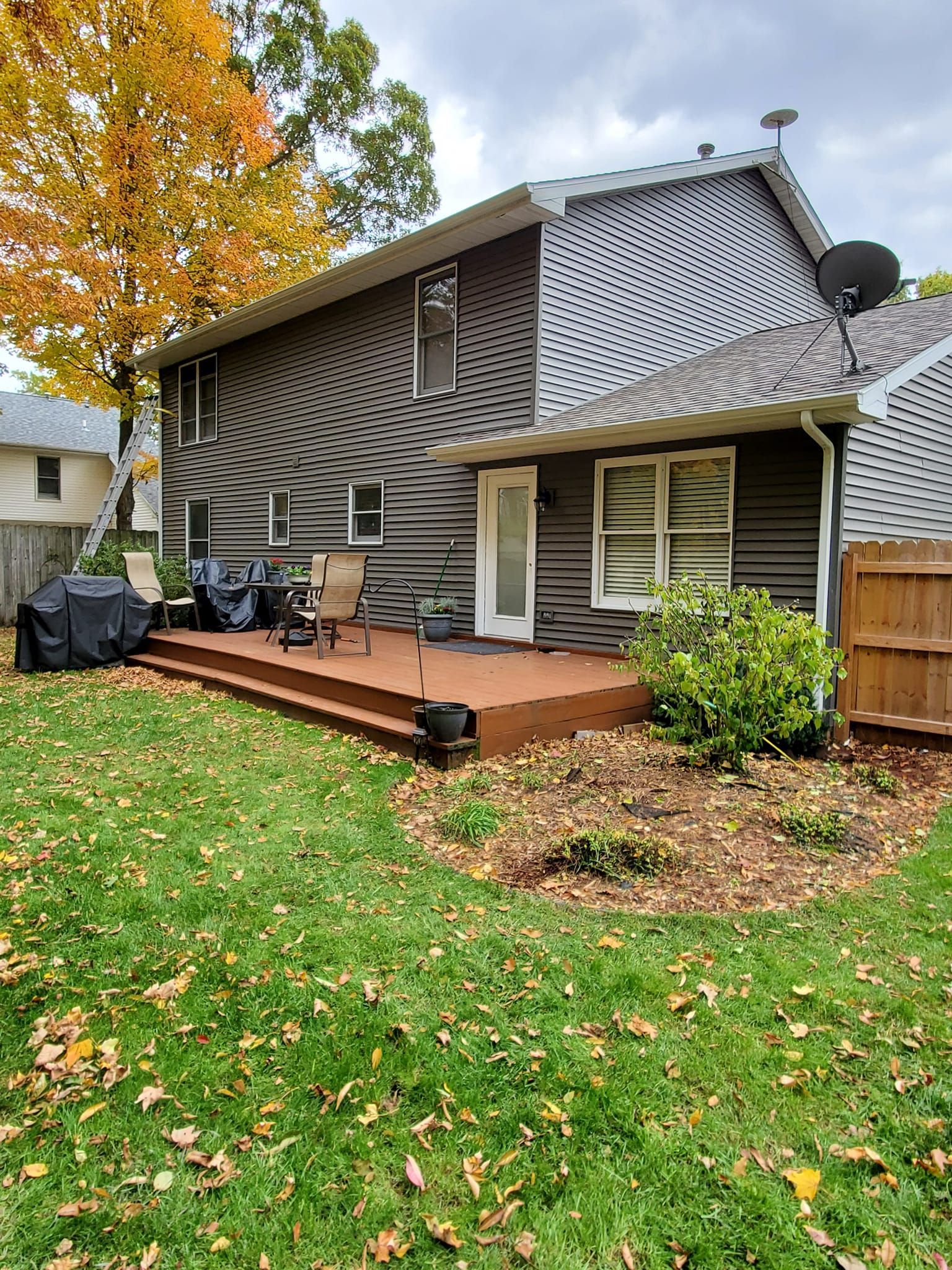 Backyard deck behind a gray house with autumn trees and fallen leaves