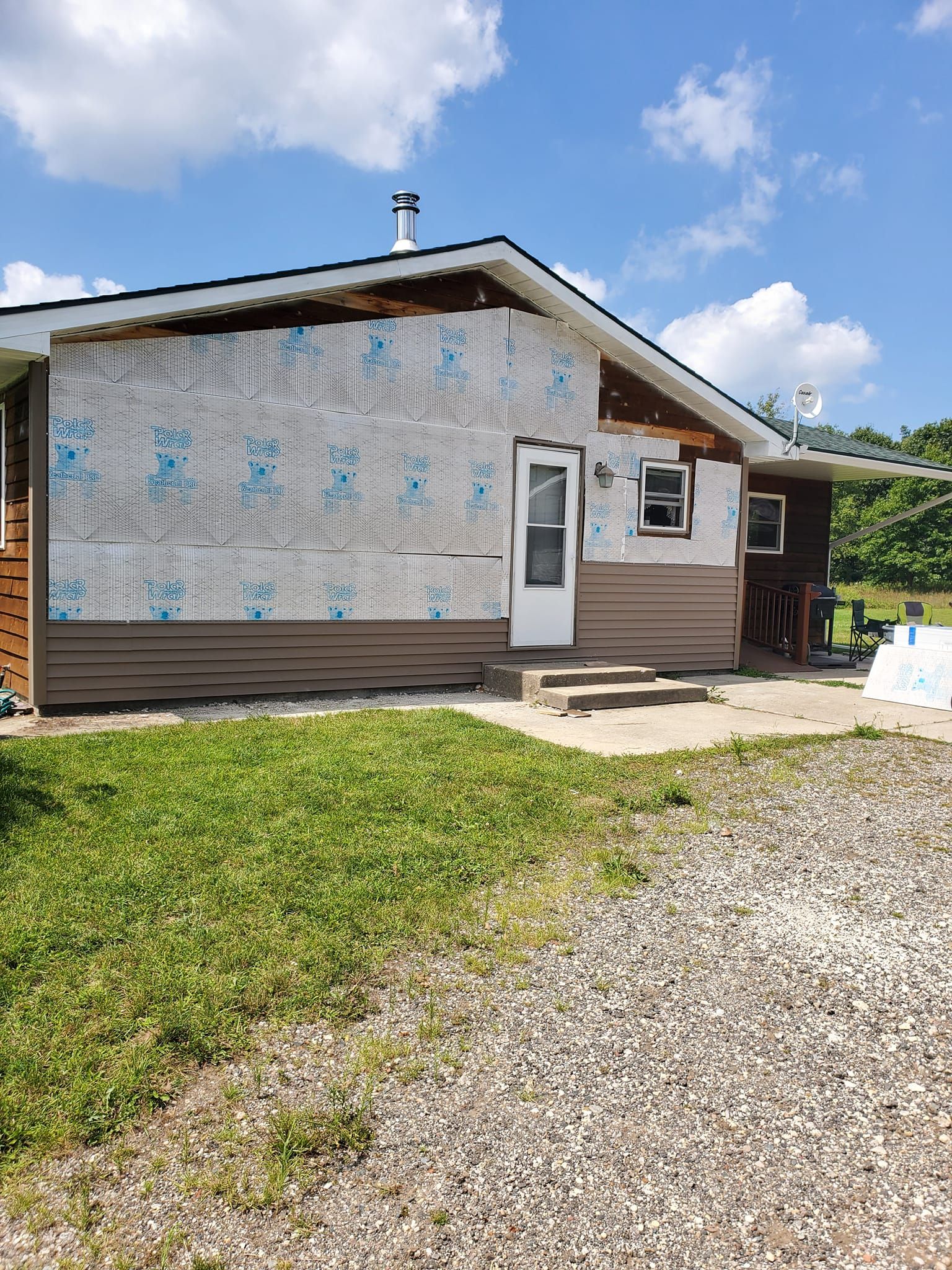 Small house with unfinished siding, gravel driveway, and grassy yard under a blue sky