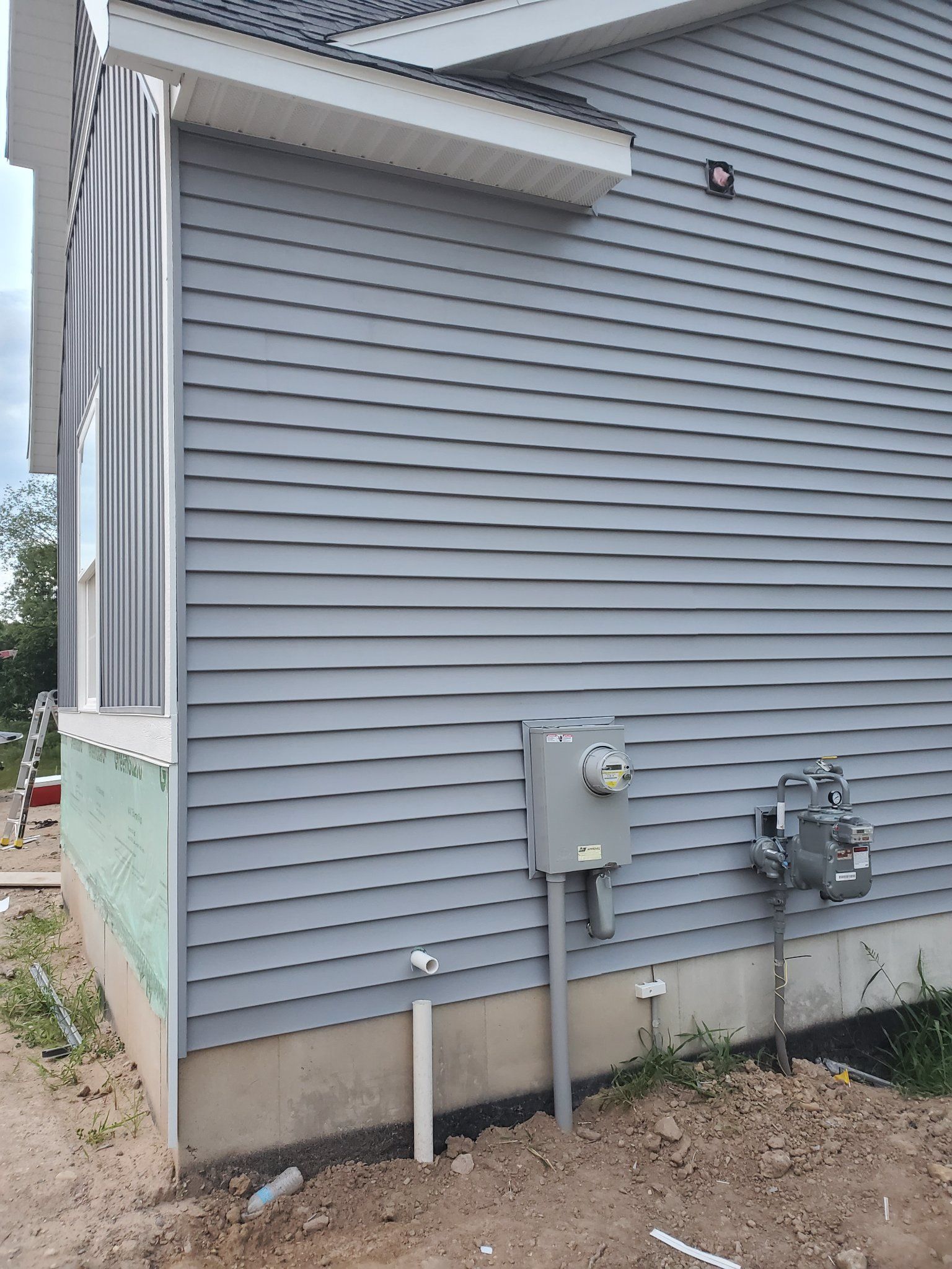 Side view of a gray house with vinyl siding, utility meters, and a small dirt yard.