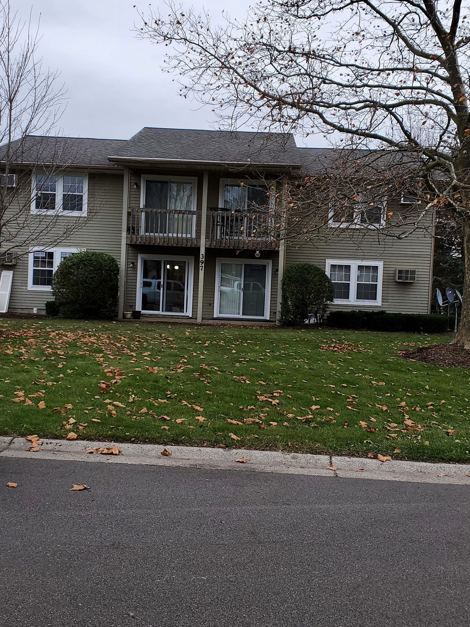 Two-story apartment building with beige siding, white trim, and a small balcony, seen across a grassy front lawn.