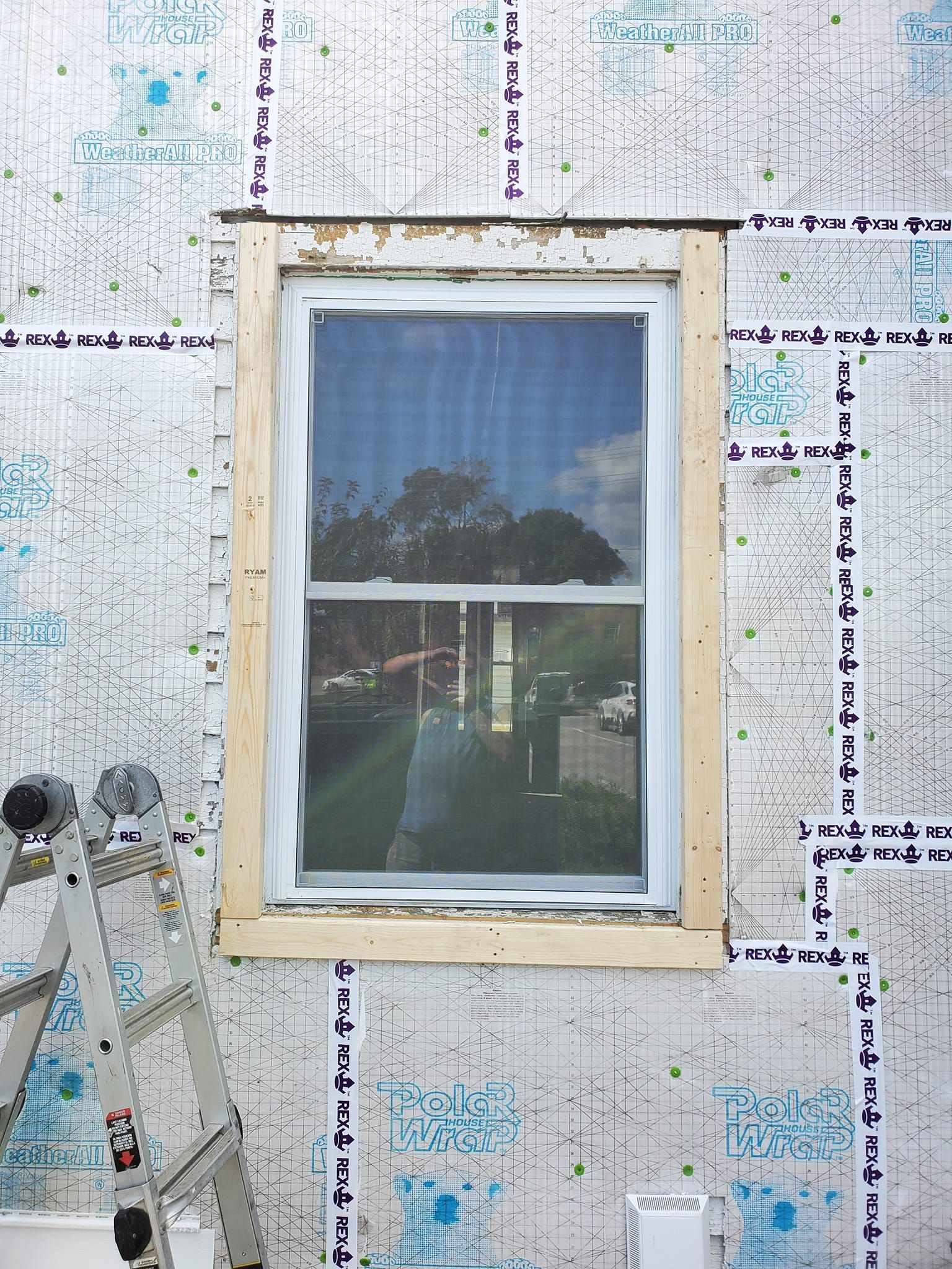 White-framed window in a sheathed exterior wall during construction, with an aluminum ladder beside it.