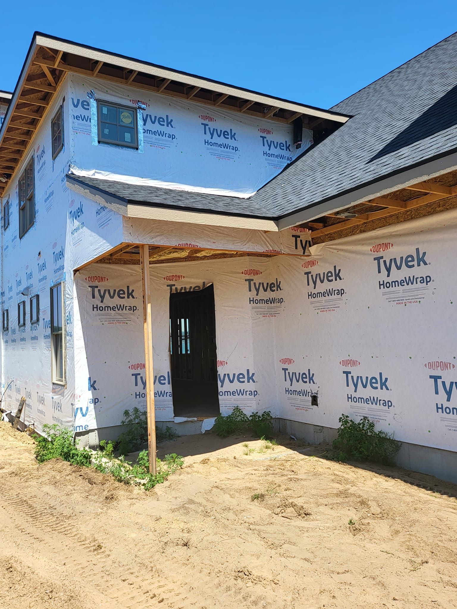 Partially built house with Tyvek wrap, stucco walls, and open doorway in a sunny dirt yard