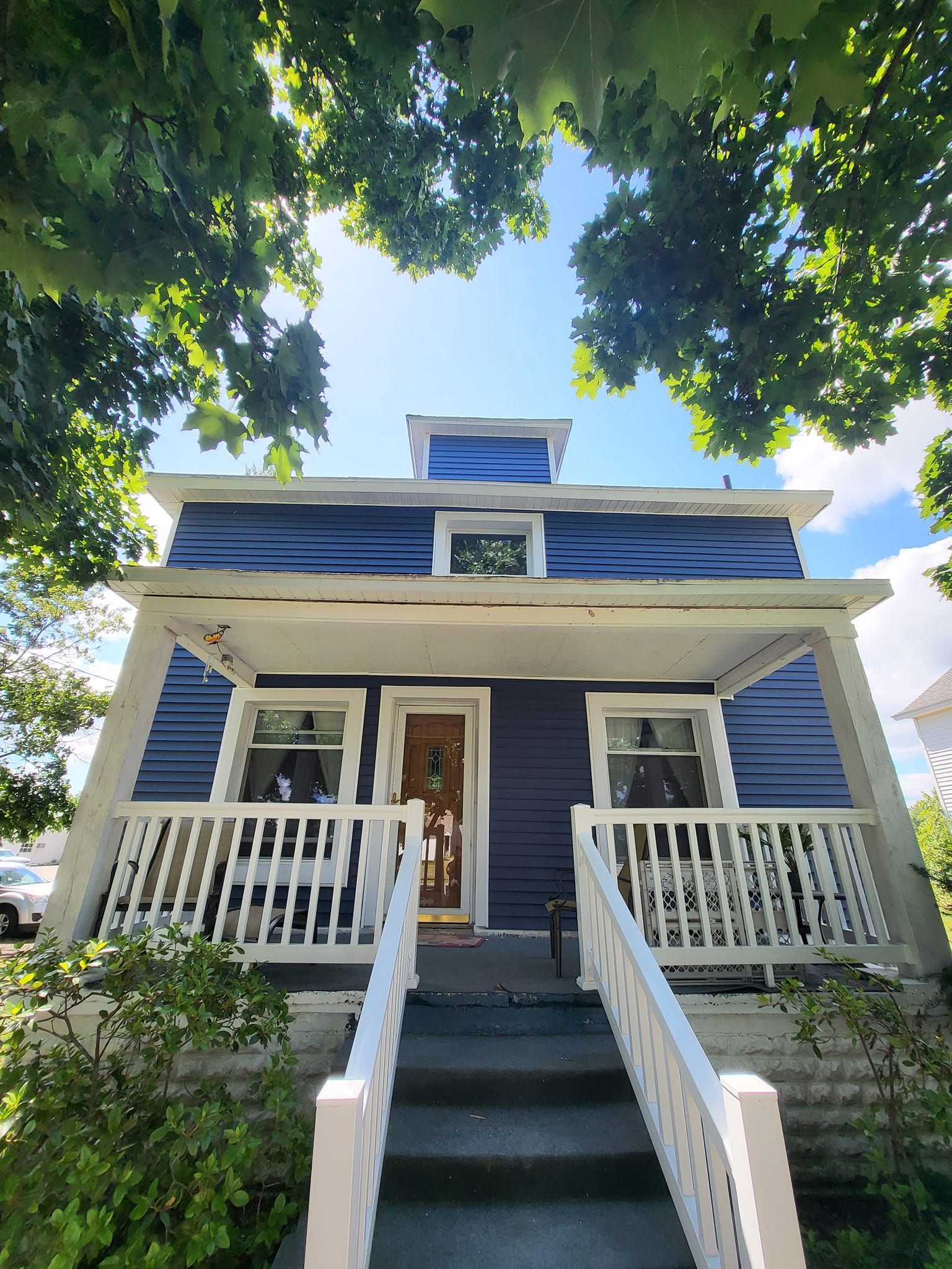 Blue house with white porch, front steps, and leafy trees overhead