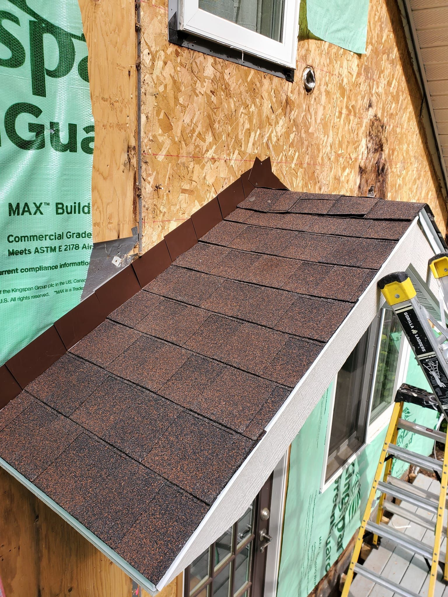 A small brown shingled roof model under construction beside green house wrap and a ladder.