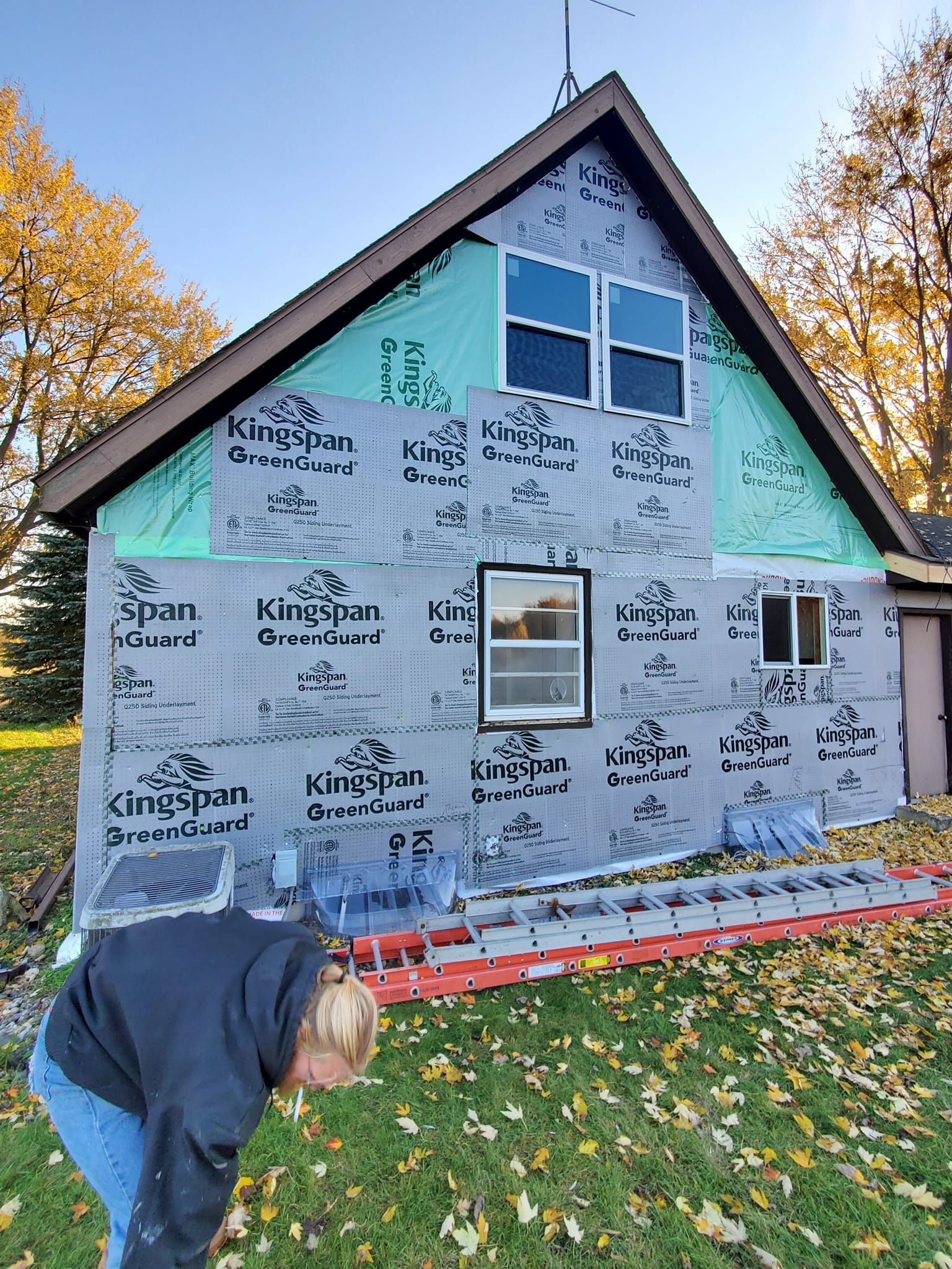 House under construction with gray house wrap and green gable trim; person bending over in foreground.