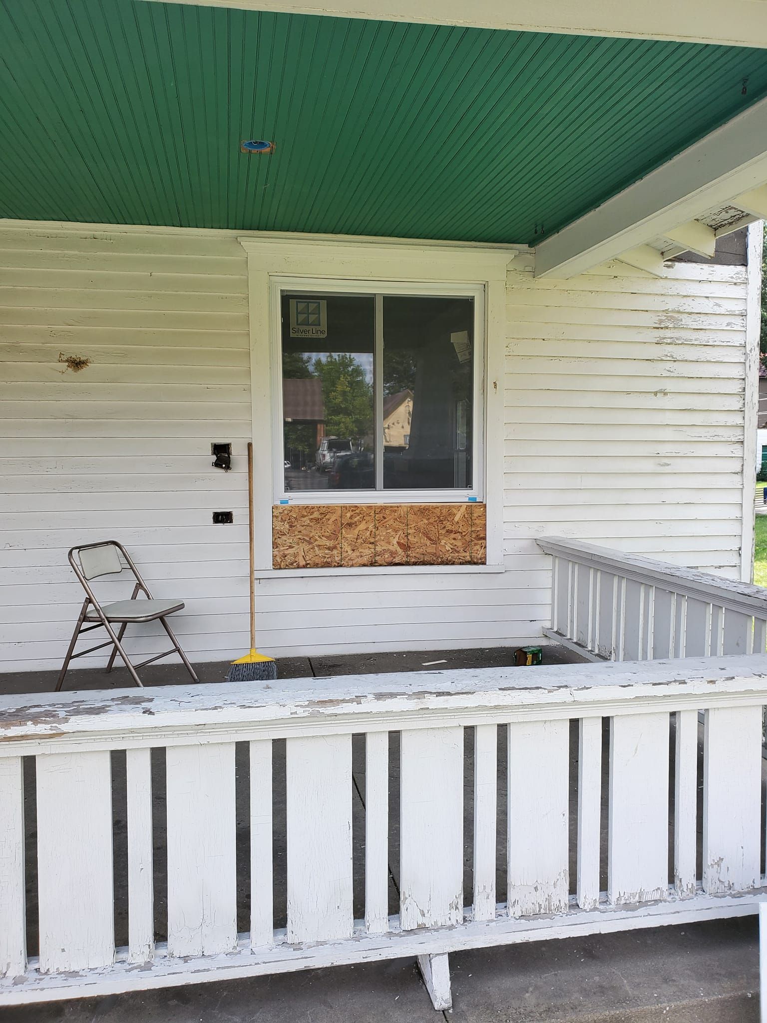 Front porch with peeling white railings, green ceiling, window, and a small chair on the left.