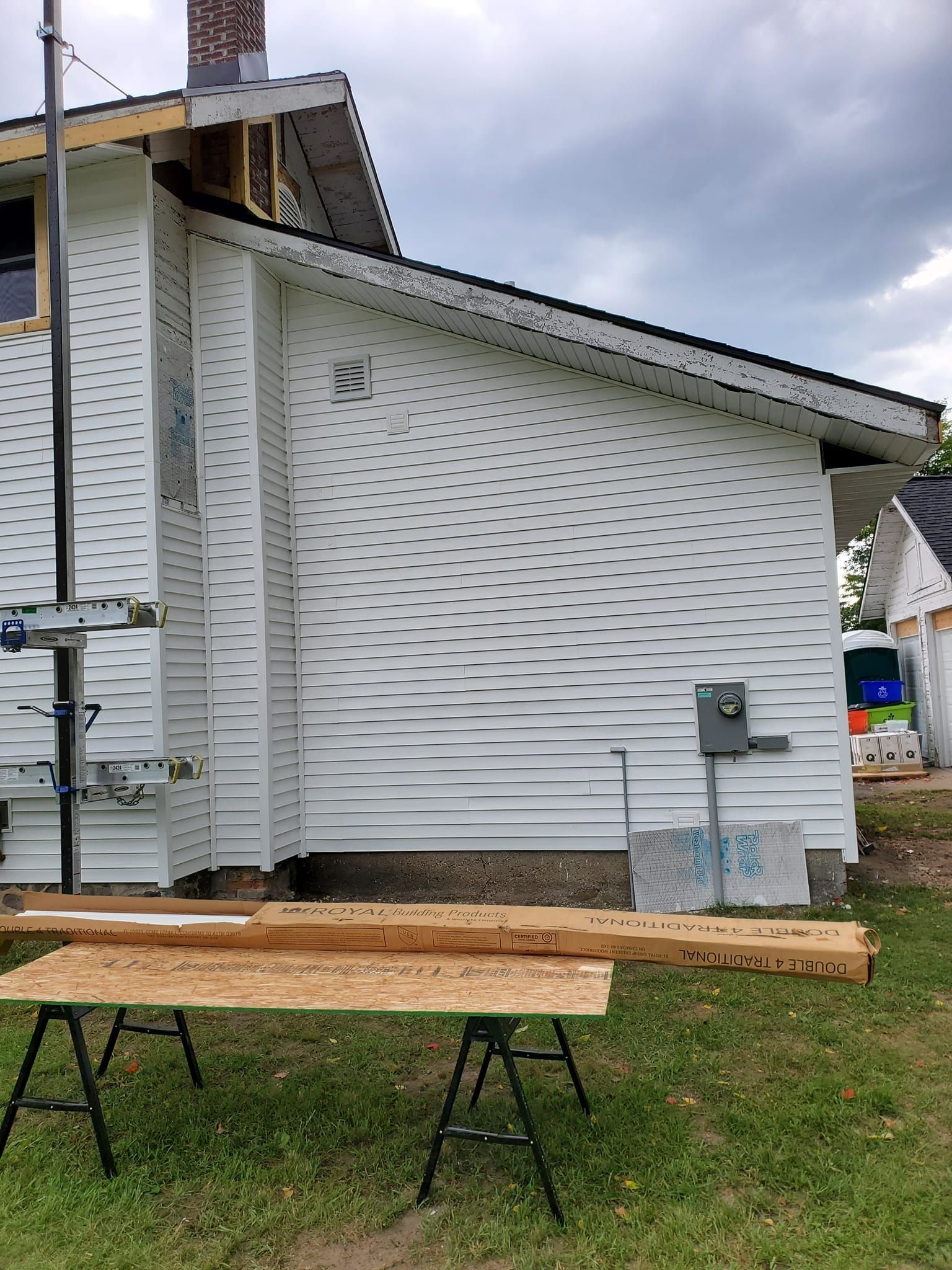 Side view of a white house with scaffolding, wooden planks on sawhorses, and a cloudy sky.