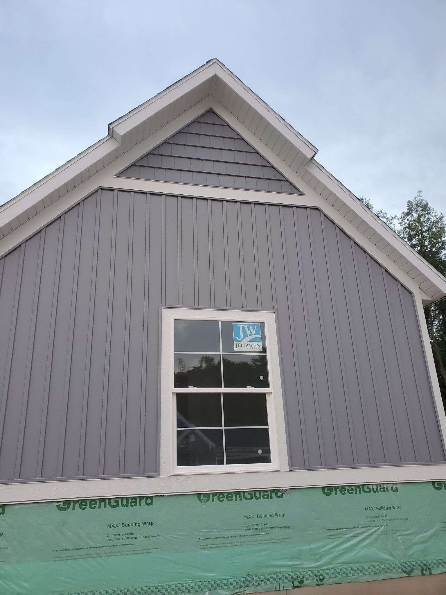 Gray house exterior with a white-trimmed window and green construction wrap at the base