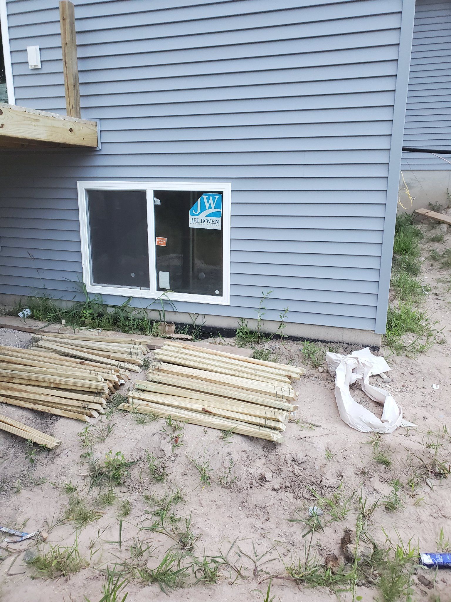 Gray house exterior with sliding window, stacked lumber, and a construction bucket on dirt ground.