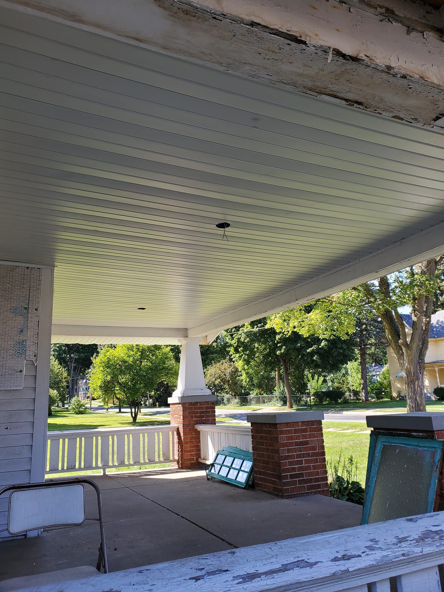 Covered porch with white ceiling, brick columns, benches, and trees in a sunlit yard.