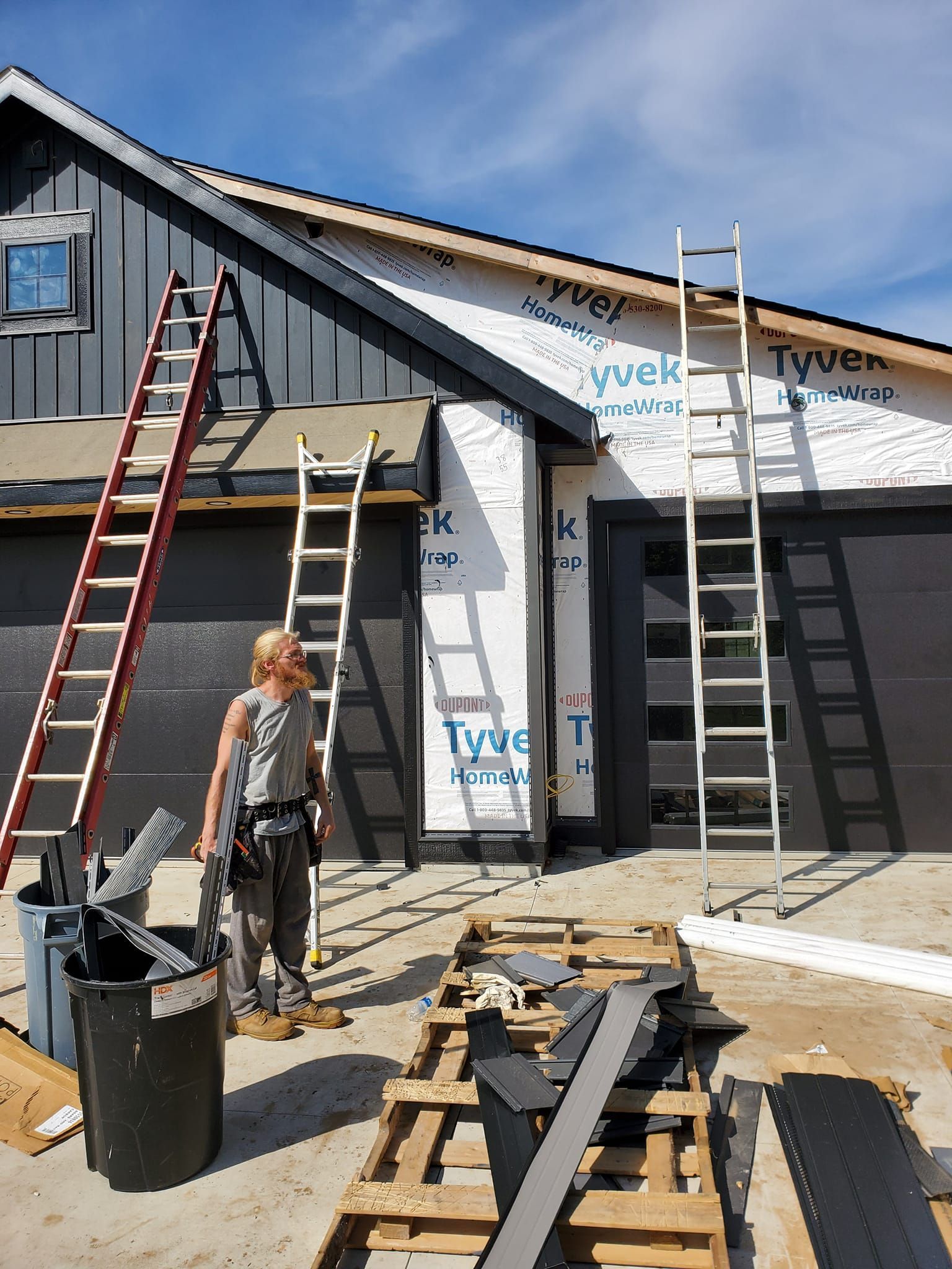 House under construction with ladders, tools, and a worker standing in front of the partially wrapped exterior.