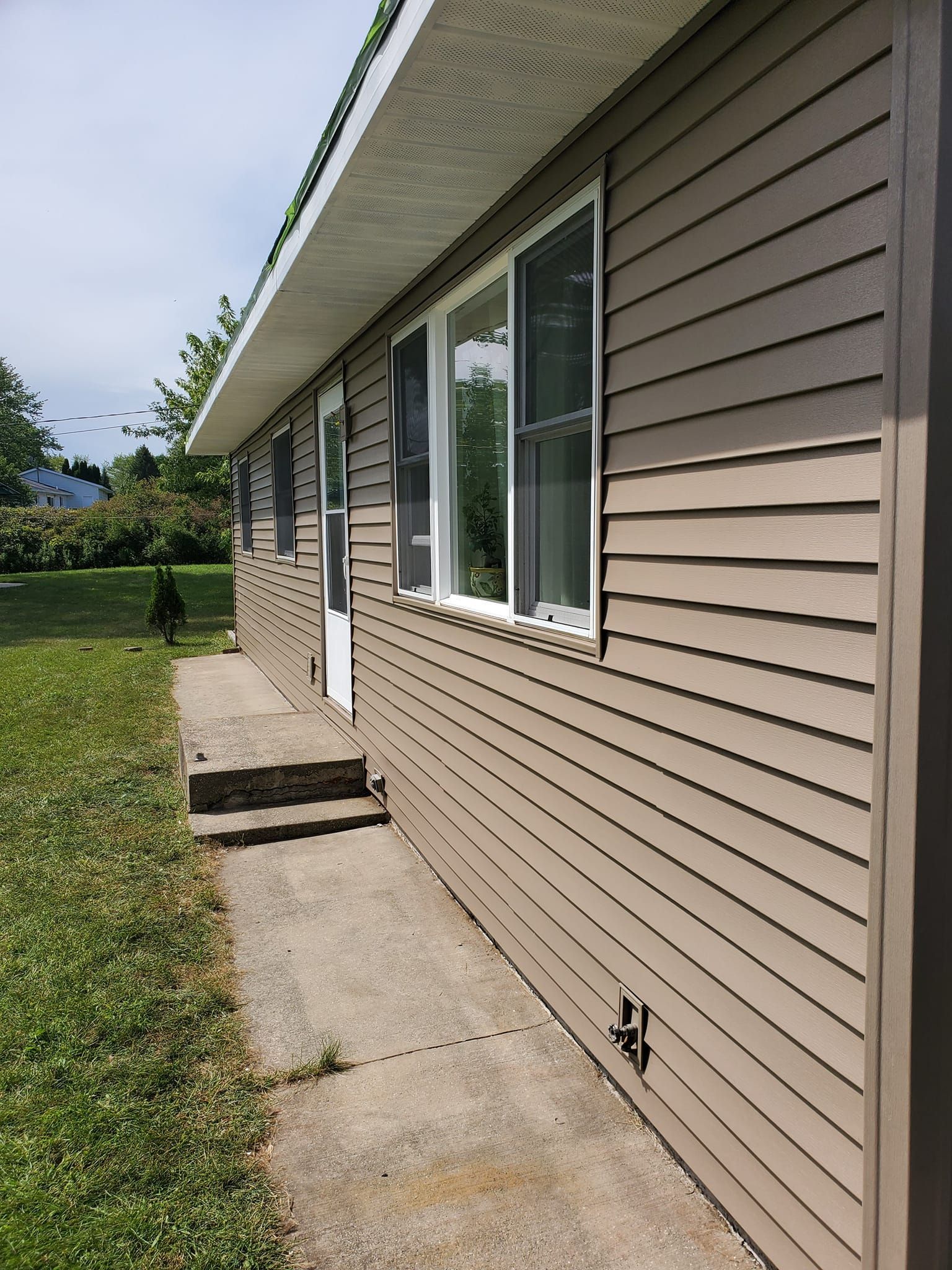 Side of beige house with windows, concrete walkway, and green lawn under an overcast sky