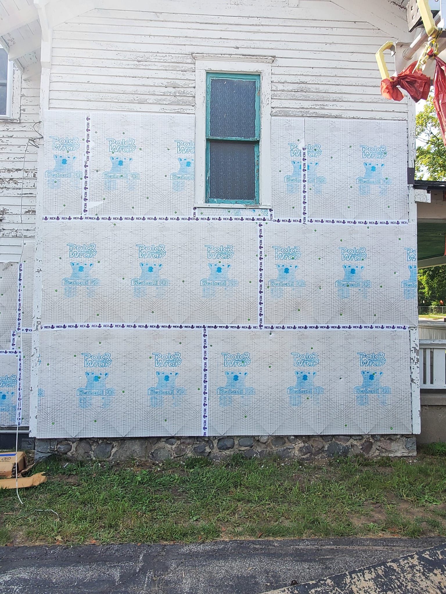 House exterior wrapped in white housewrap with a central window and visible blue markings, above grass and sidewalk