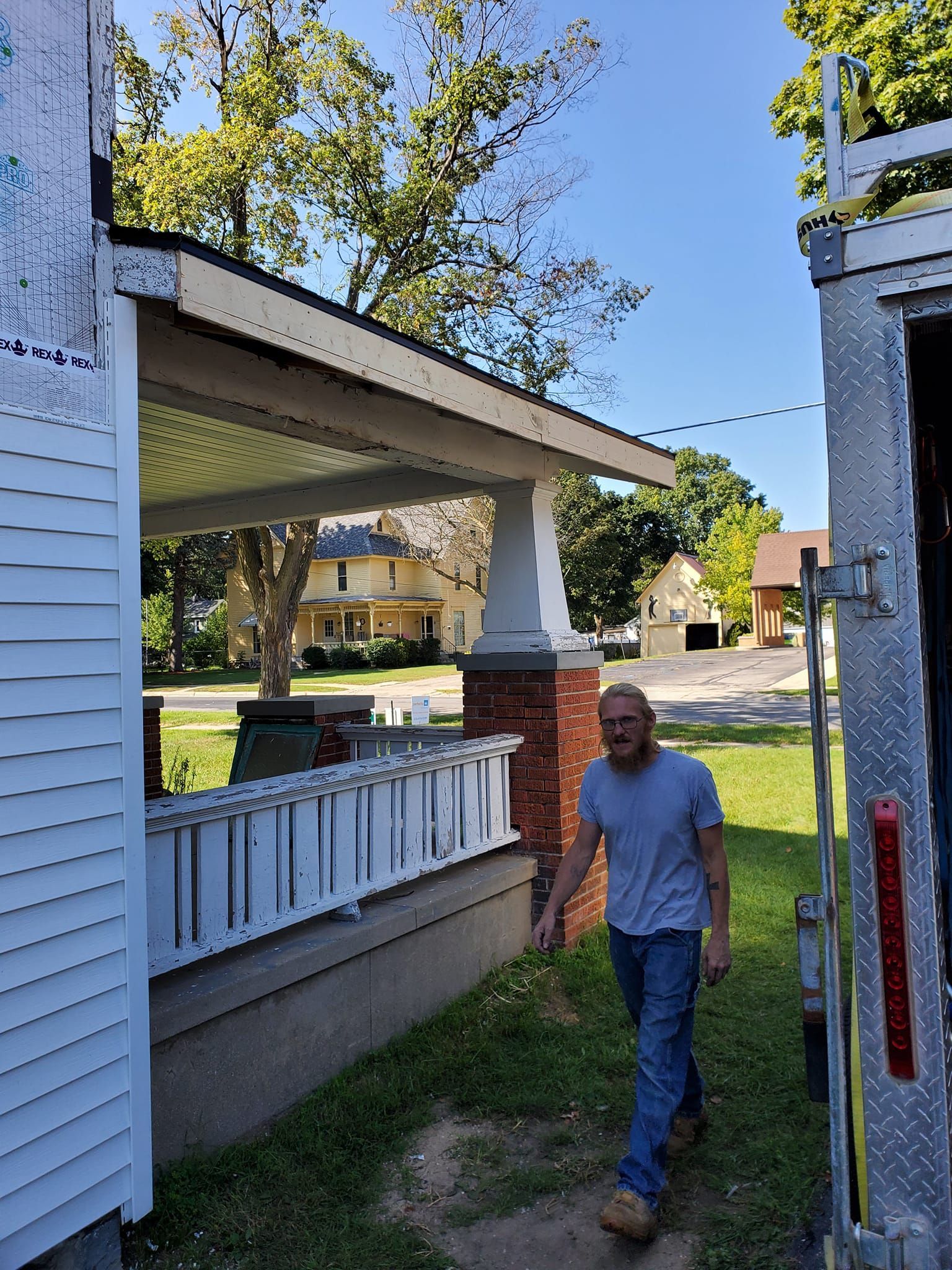 Man walking beside a white porch and brick pillar in a sunny yard with trees and neighboring houses.