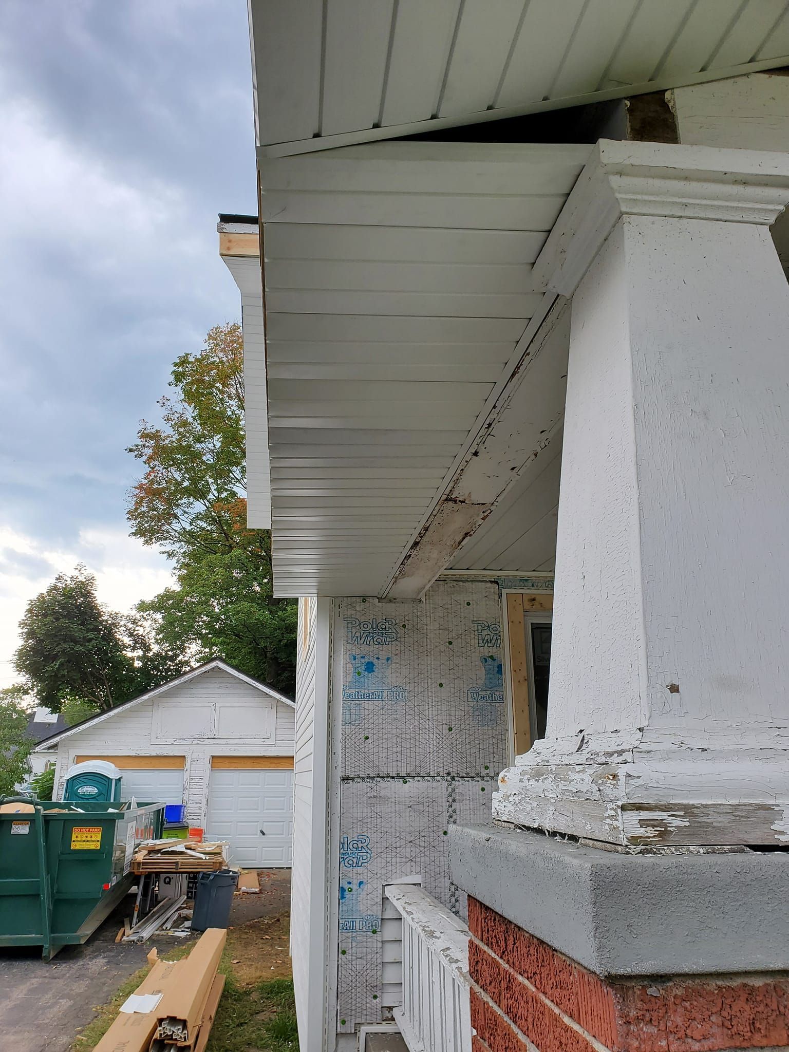 Side of a weathered house with white trim, peeling paint, and a garage, with a green dumpster and trailer nearby.
