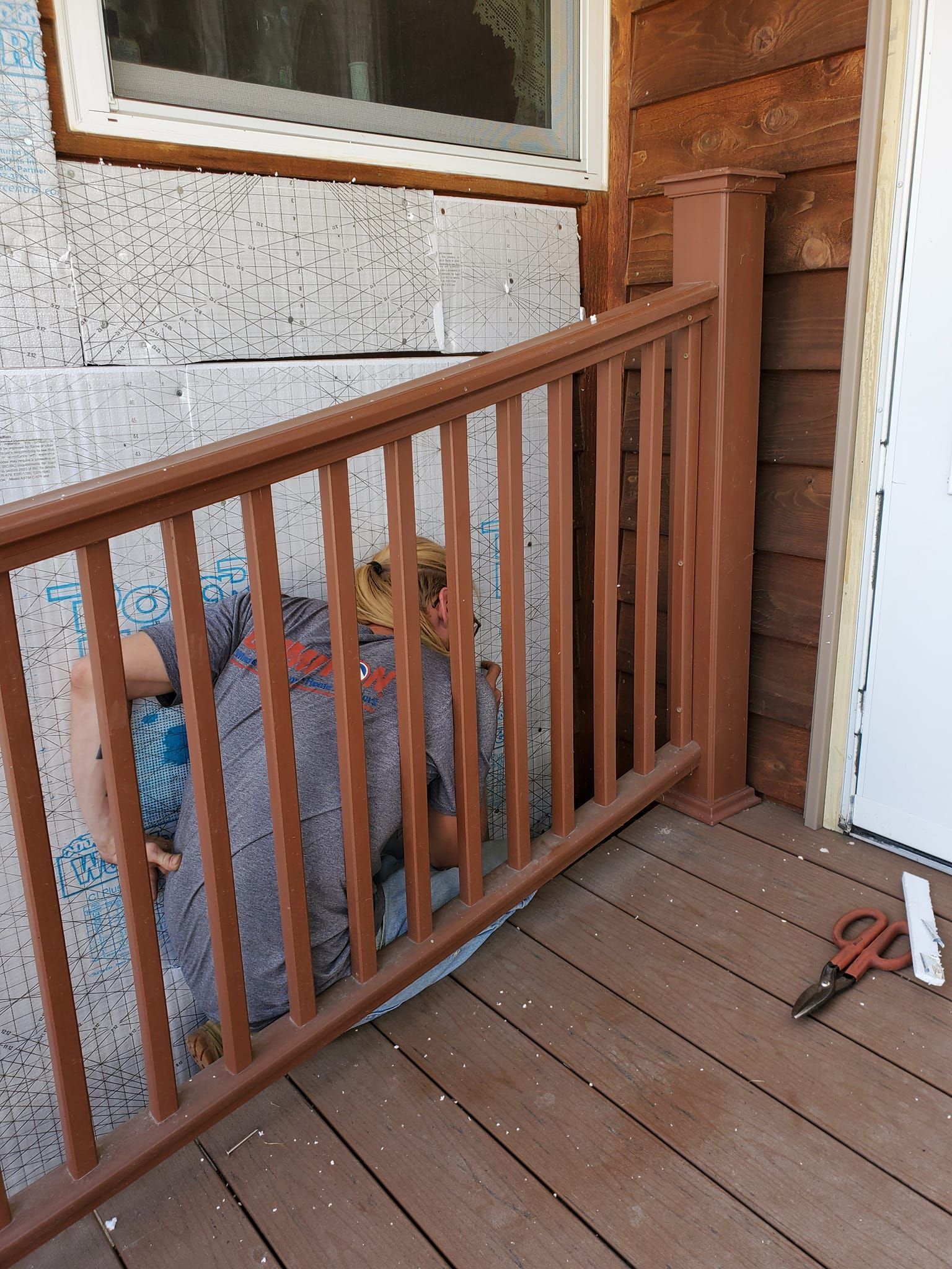 Wooden porch railing beside a window and white door, with a red tool on the floorboards.