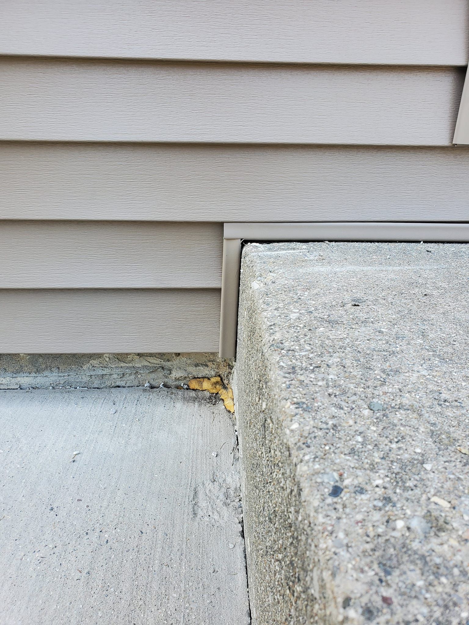 Exterior house wall with gray siding and concrete foundation corner, showing a small gap and debris.