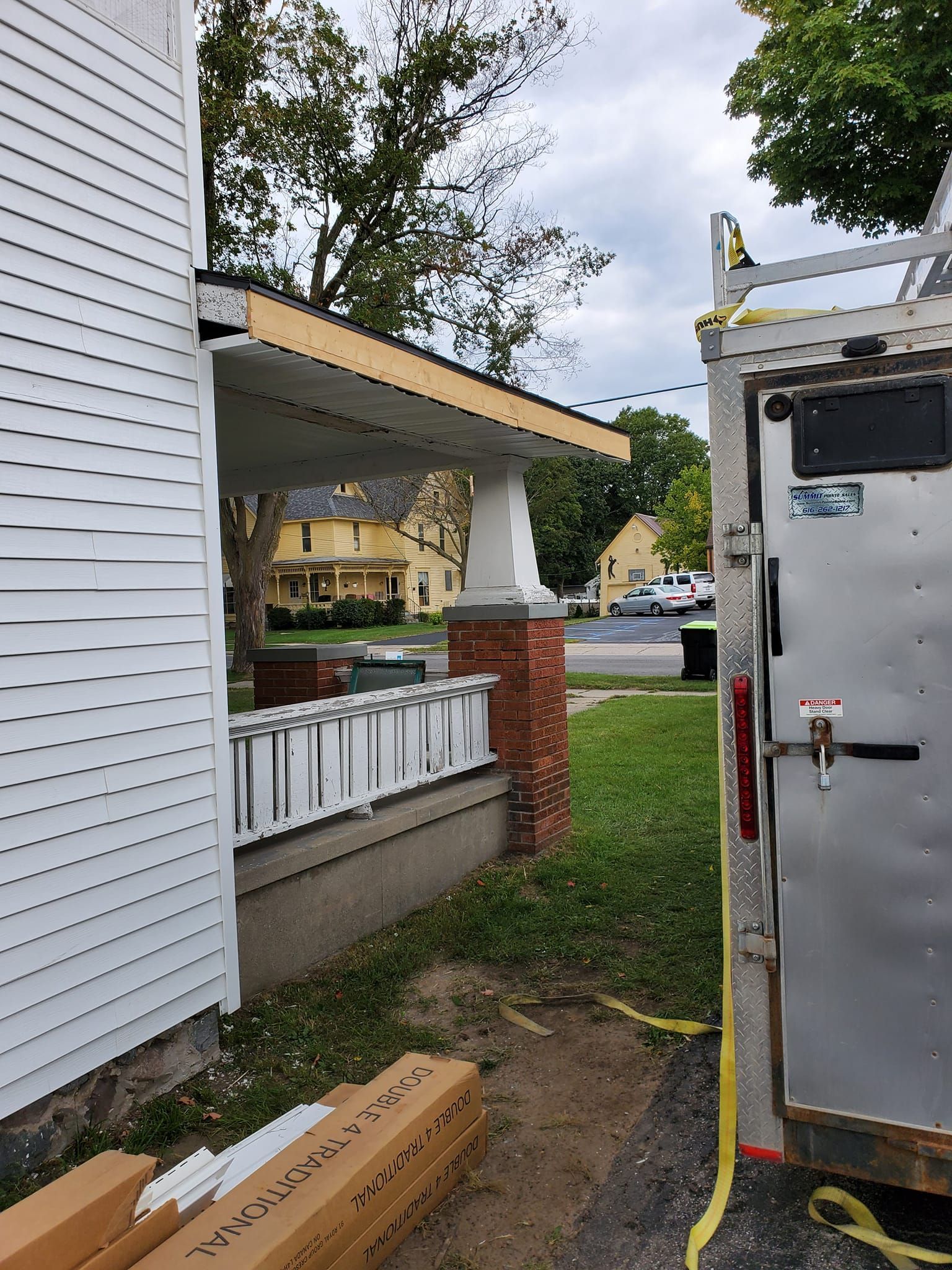 House siding and porch beside a white trailer with a yellow hose in a narrow driveway.