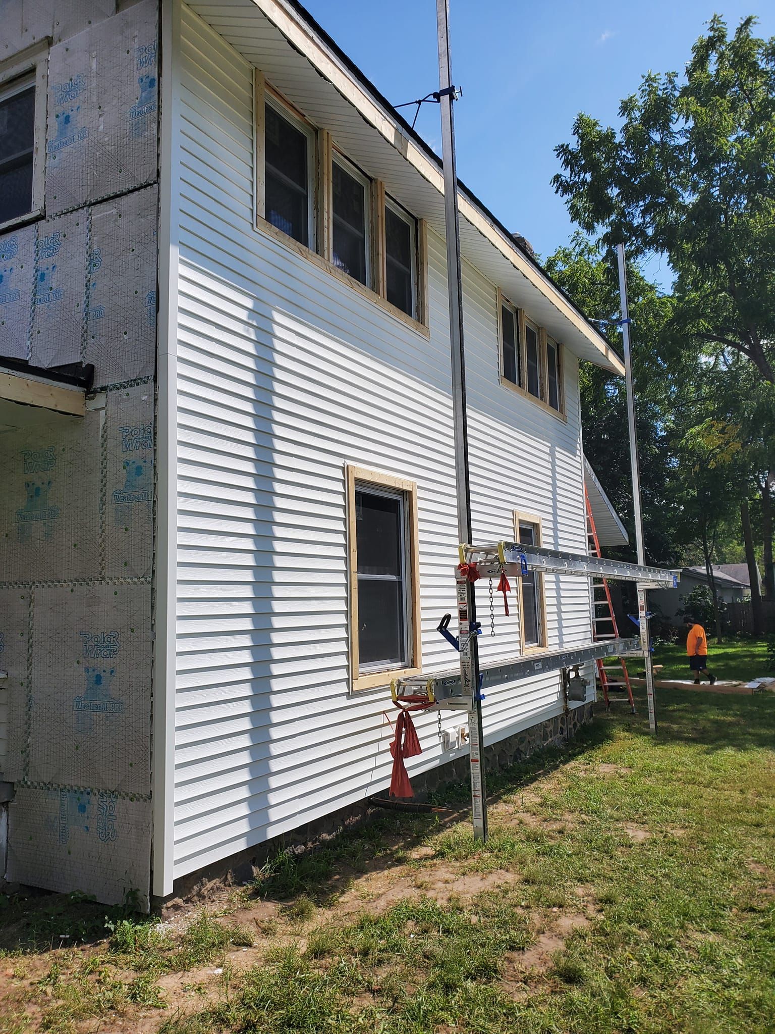 White two-story house exterior with scaffolding and red pipes beside a grassy yard