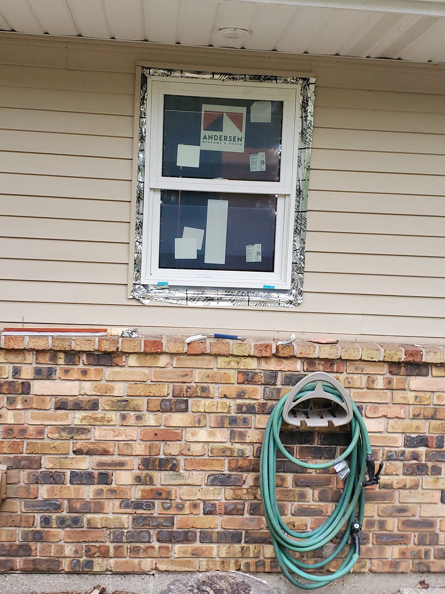 House exterior with two windows, brick wall, and a coiled green garden hose mounted below.