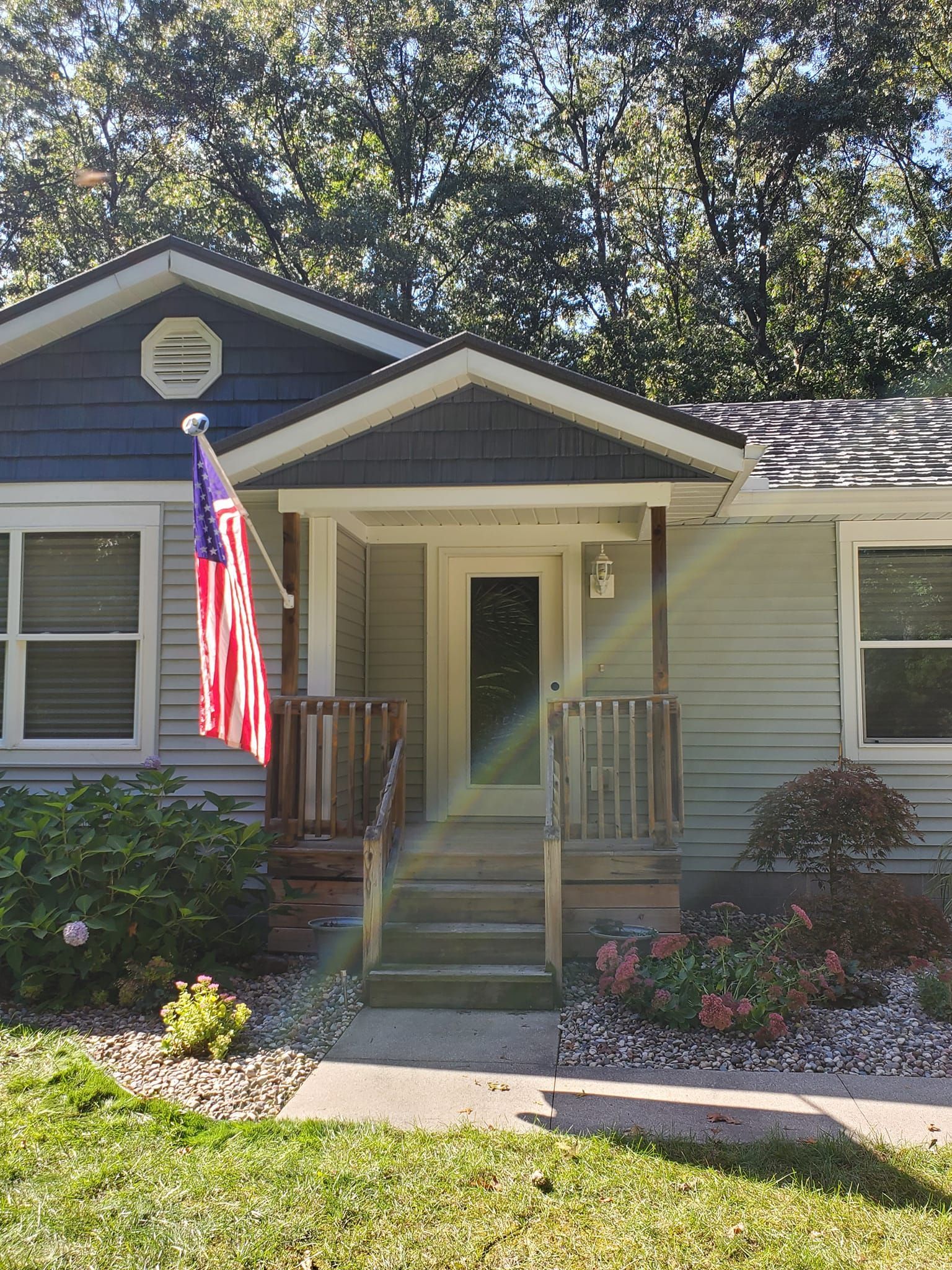 Small house with a front porch, American flag, and flower garden in bright daylight.