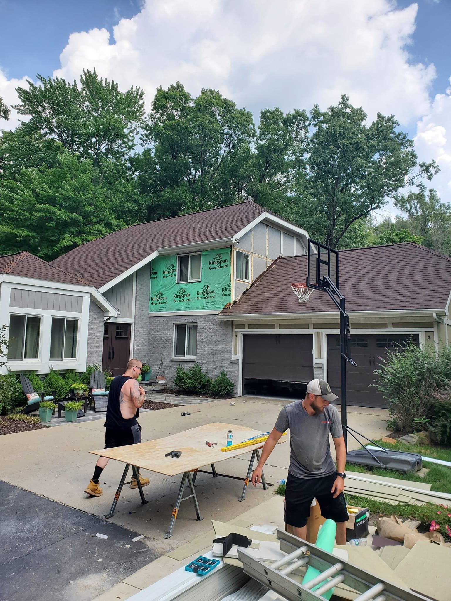 People setting up folding tables and a basketball hoop in a driveway outside a house