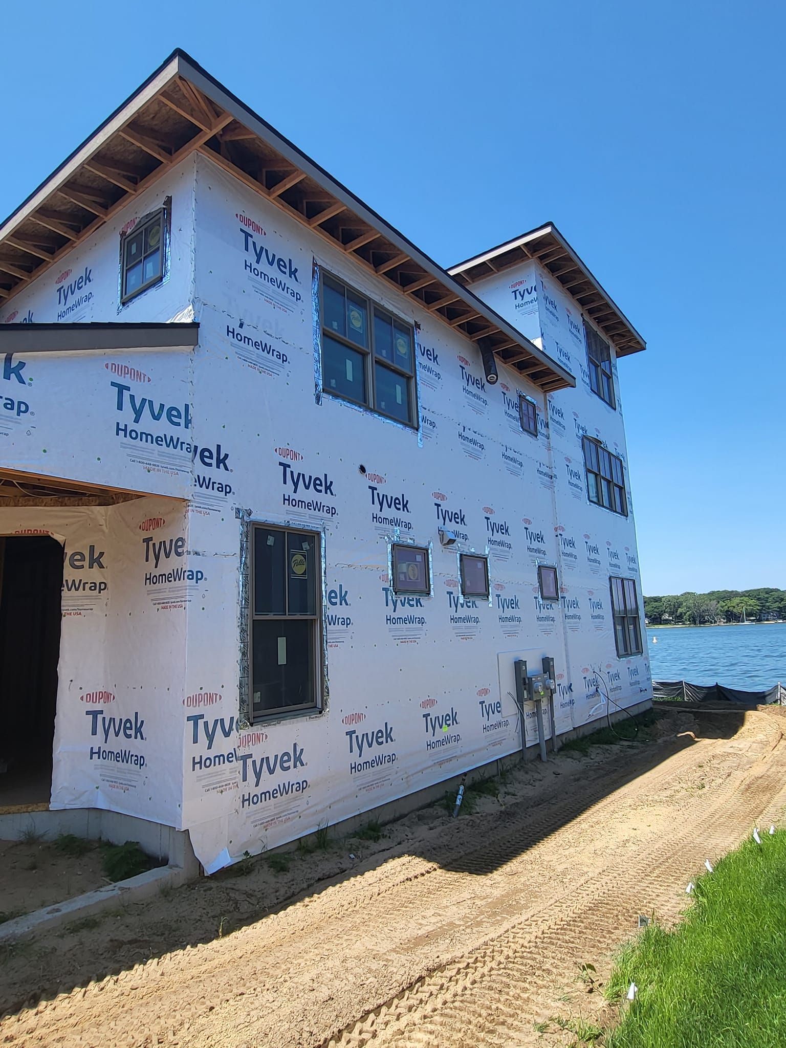 Two-story house wrapped in white construction wrap beside a dirt path and water under a blue sky