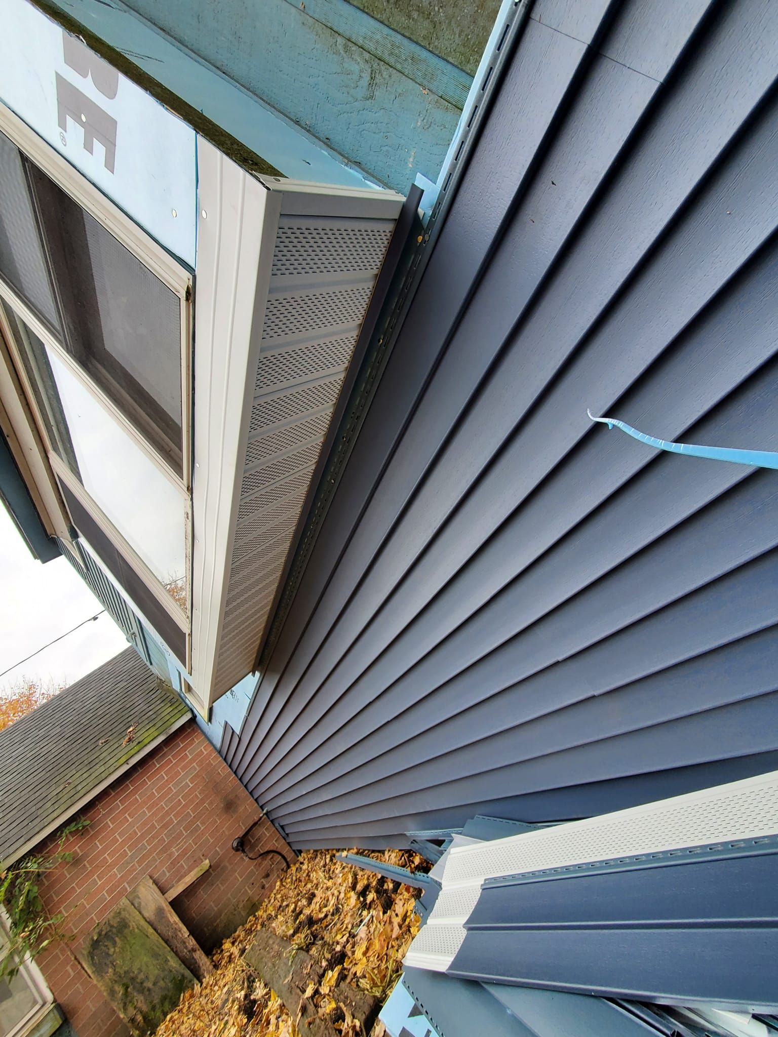 Tilted exterior view of a house corner with blue siding, windows, roof edge, and brick and leaves below