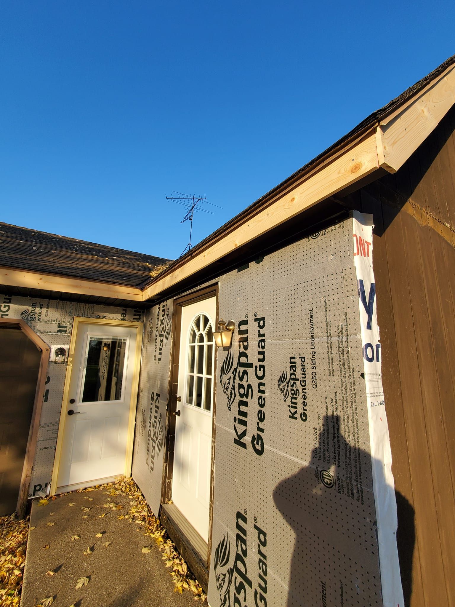 Damaged house exterior with exposed wall wrap, torn siding, and blue sky overhead