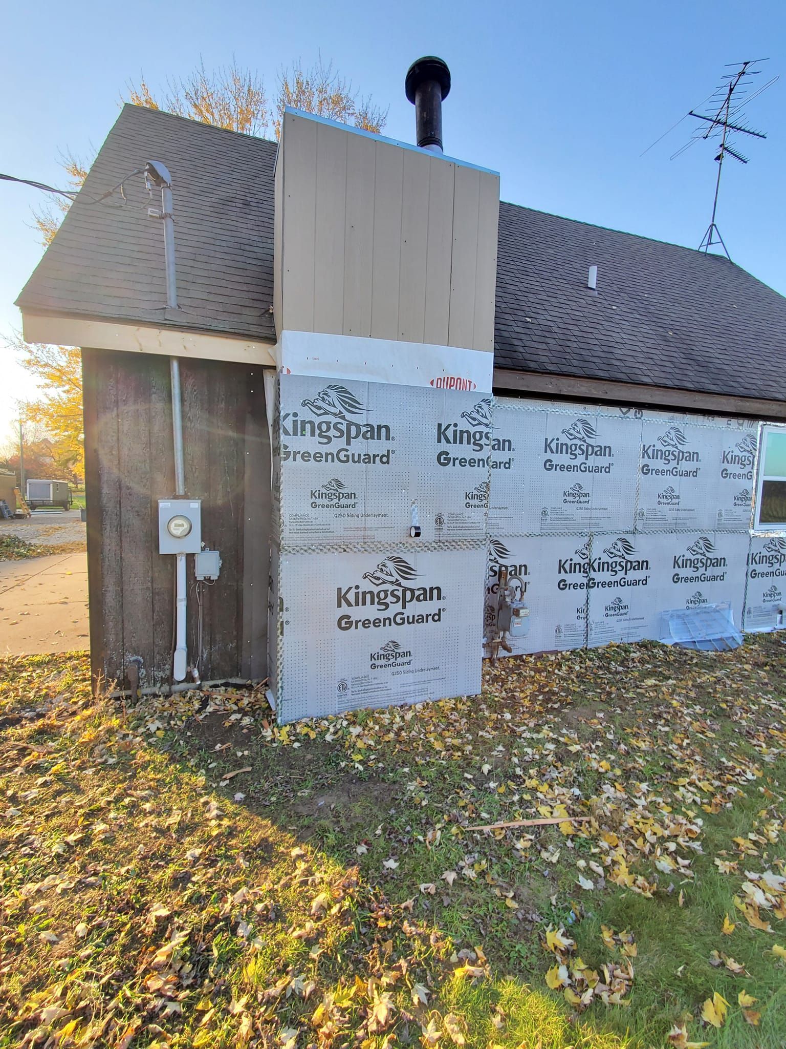 House exterior under renovation, wrapped in white siding panels and housewrap, with a chimney and fallen leaves in front.