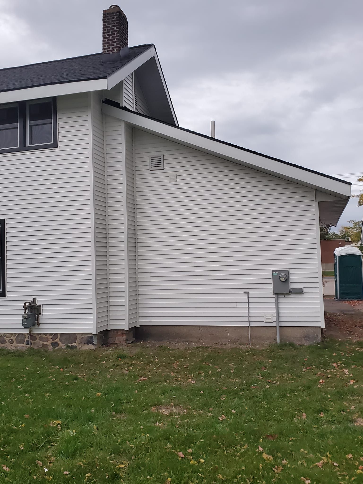 Side view of a white house with gray roof, chimney, utility meter, and grass yard.
